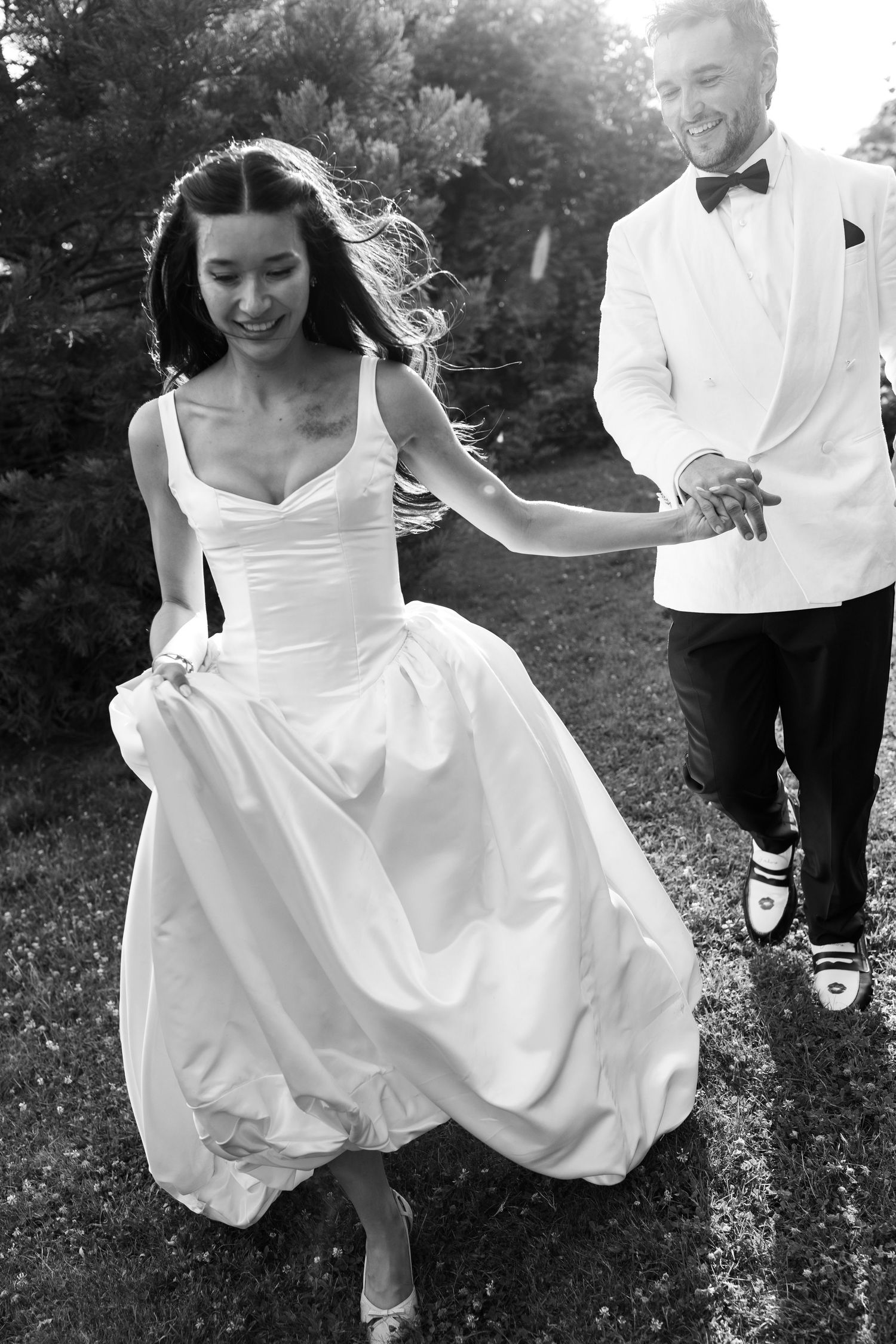 Black and white photo of a joyful bride in a flowing gown and groom in white tuxedo running hand in hand outdoors.
