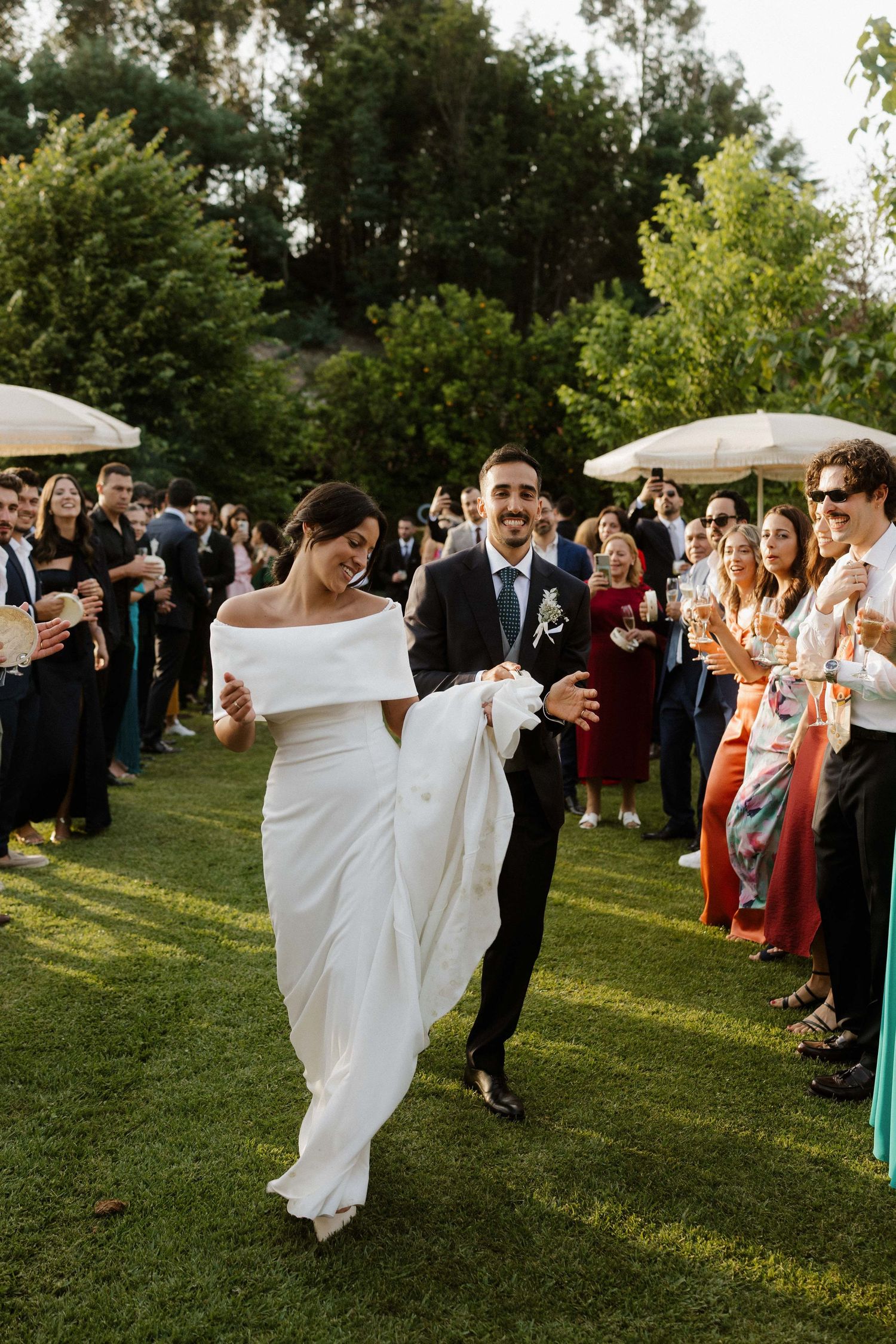 Radiant bride and groom walking through cheering guests on a sunny green lawn at outdoor wedding.