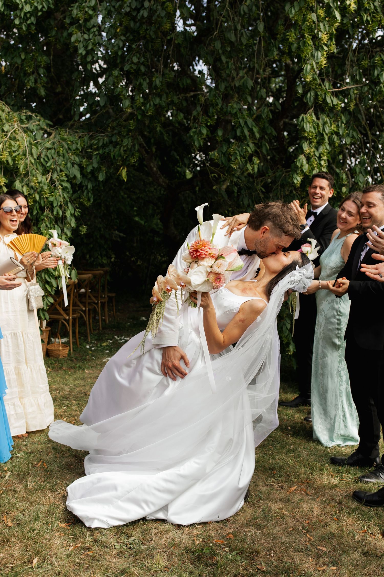Groom dips bride for a romantic kiss at outdoor wedding ceremony while guests celebrate around them.