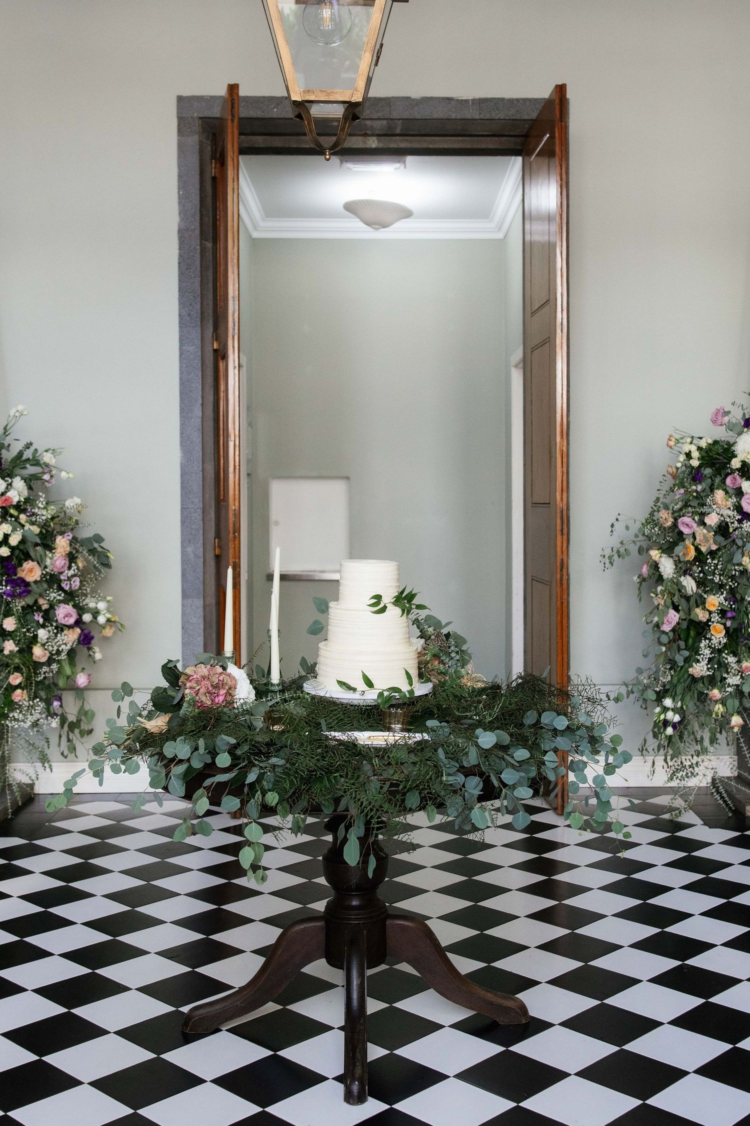 Elegant three-tier white wedding cake on a wooden table with lush floral arrangements on a checkered floor.