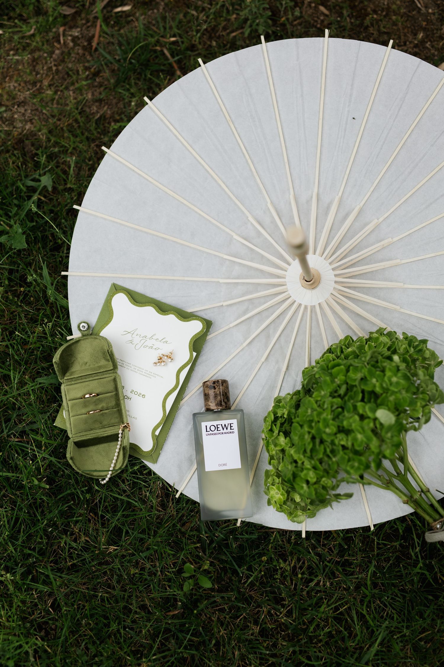 Bridal flat lay featuring Loewe perfume, green ring box, wedding invitation, and hydrangeas beside white parasol on grass.