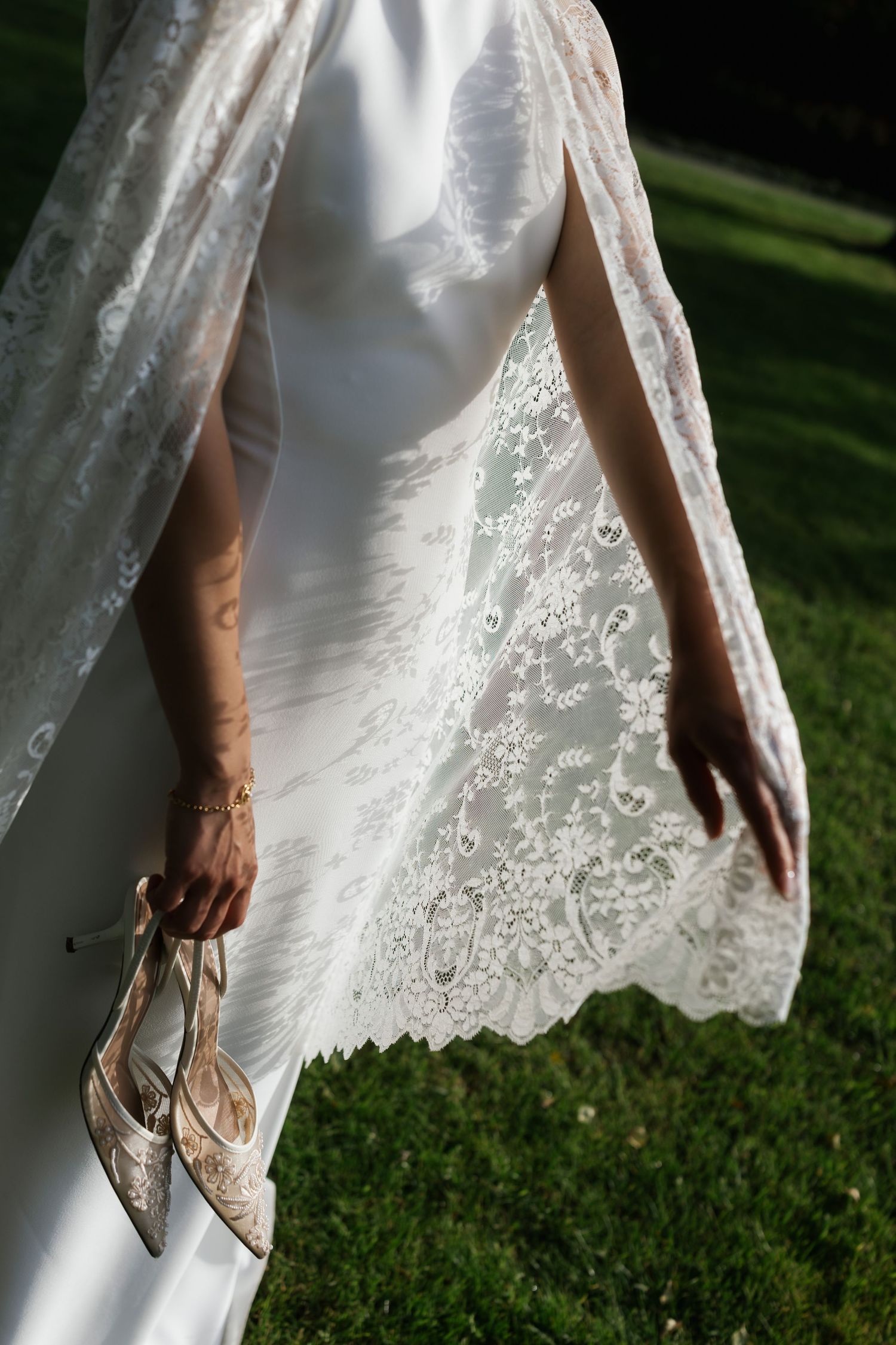 Bride in white gown with lace cape holds strappy heels while walking on green grass in sunlight.