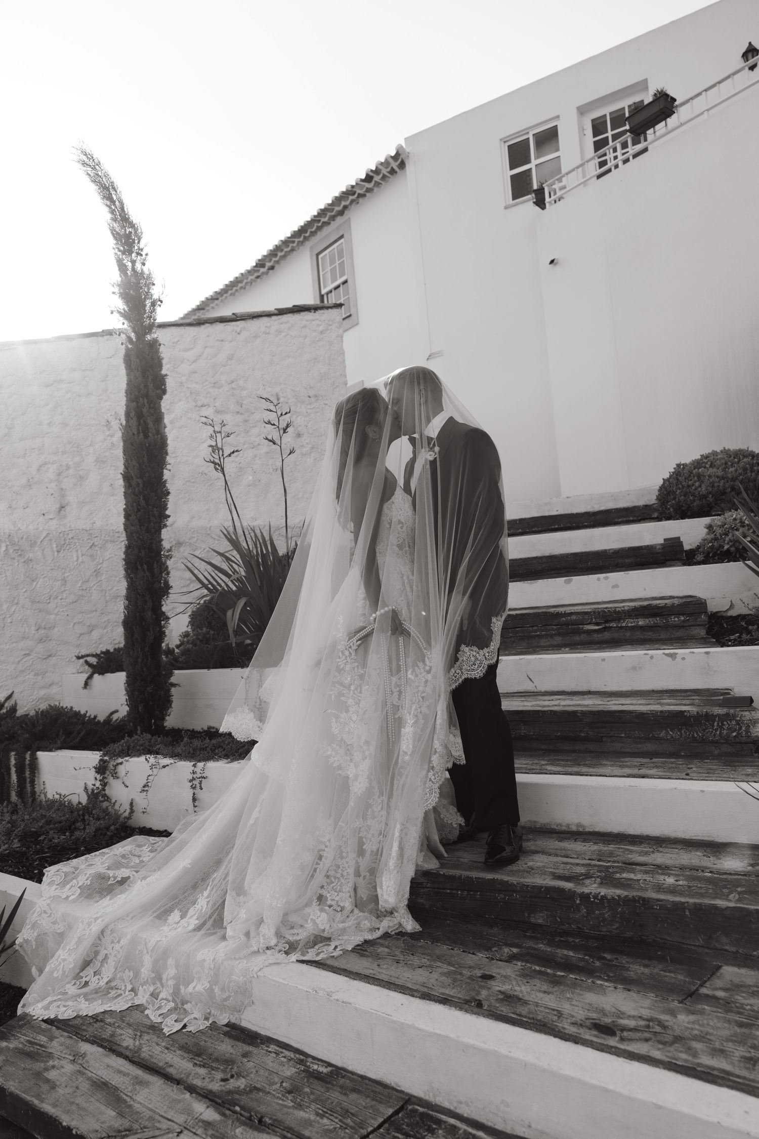 Bride and groom share a kiss on rustic wooden steps with lace cathedral veil trailing, black and white photo.