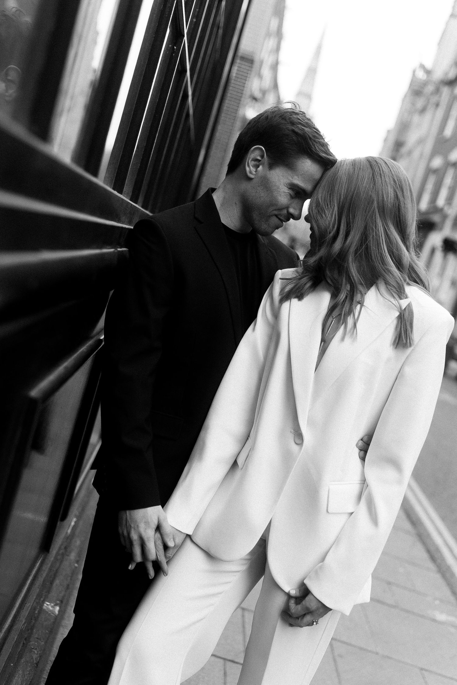 Black and white romantic photo of couple in white and black suits embracing on city street.