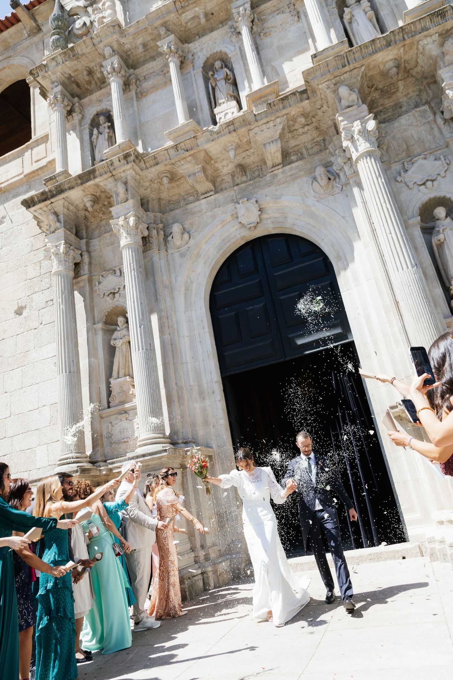 Newlyweds exit grand baroque church entrance as guests celebrate with confetti shower and champagne spray.
