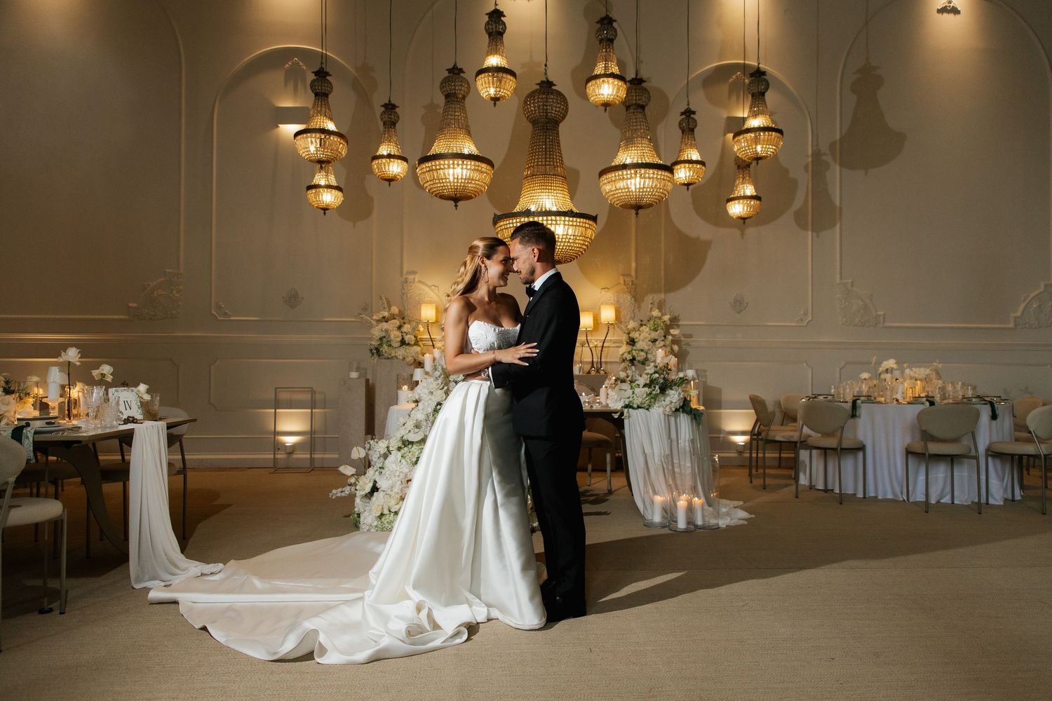 Bride and groom share romantic embrace in ornate ballroom with cascading chandeliers and white floral arrangements.