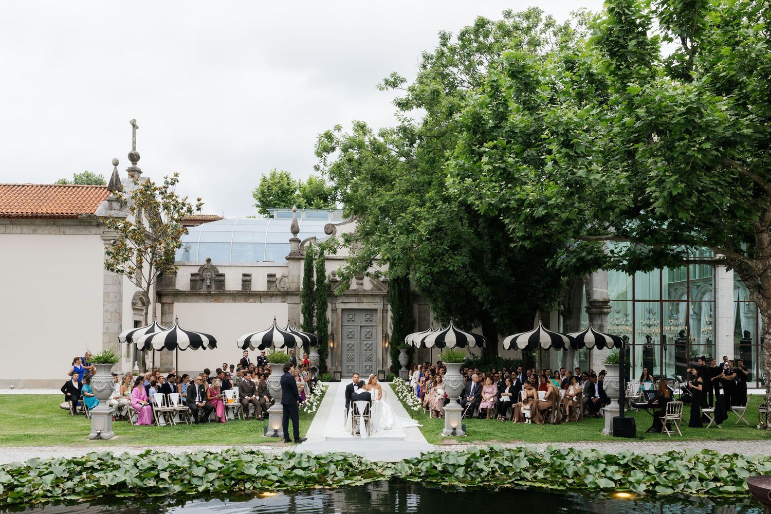 Aerial view of outdoor wedding ceremony beside lily pond with black and white striped umbrellas and lush gardens.