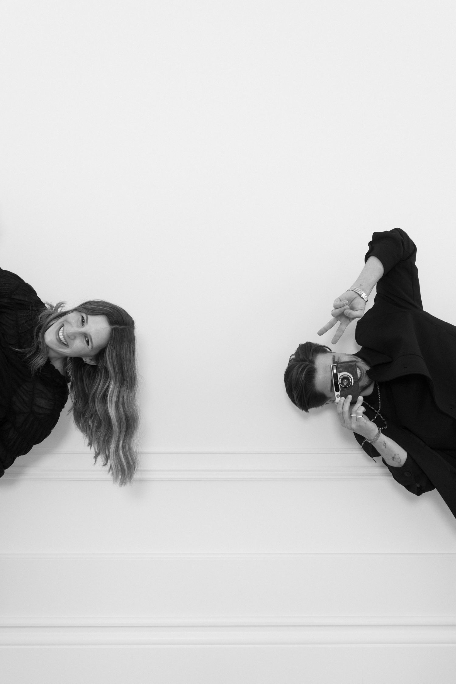 Black and white photo of two people in black outfits, one smiling and one holding a camera, mirrored.
