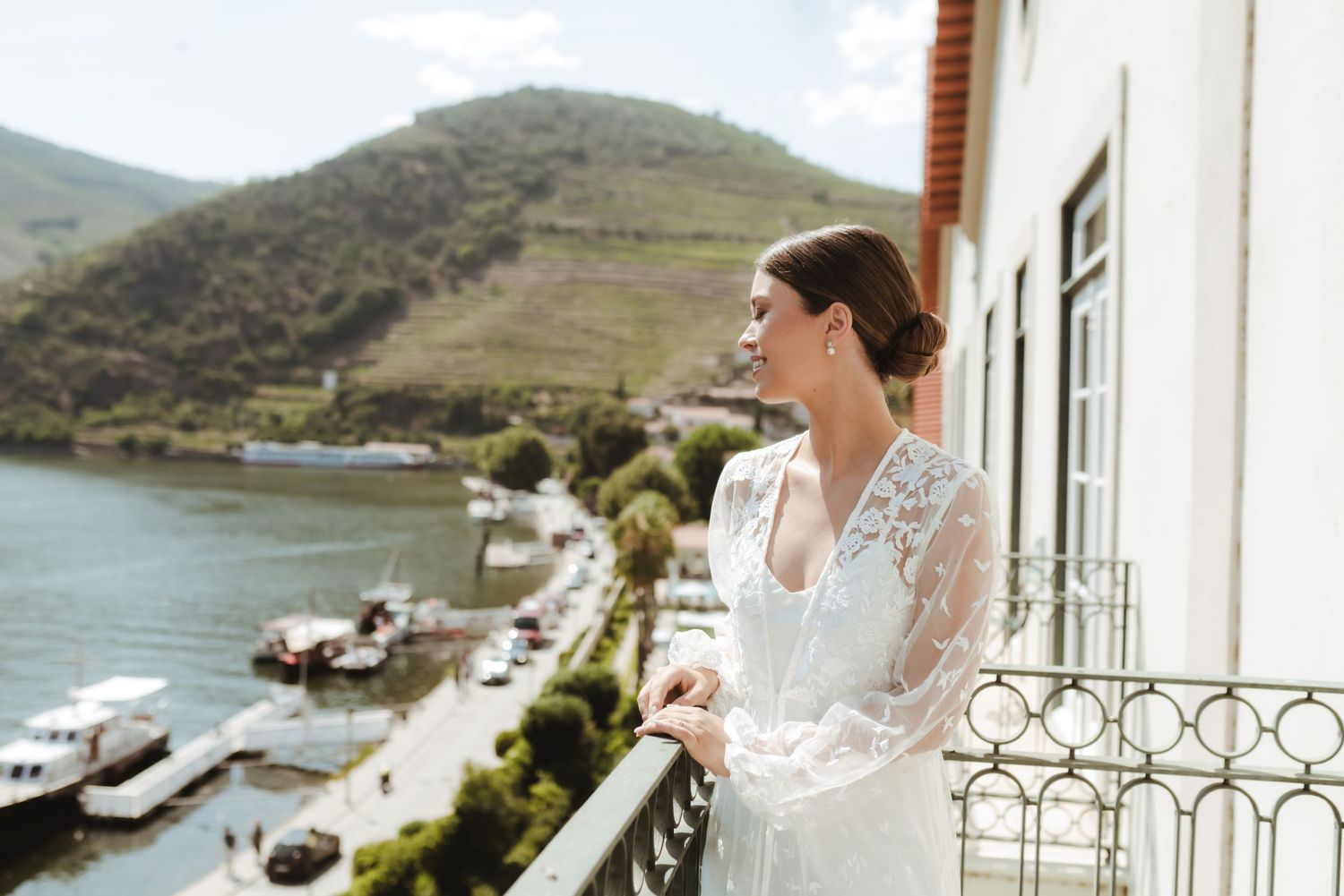 Bride in lace wedding gown standing on balcony overlooking scenic river valley and green hillside landscape.