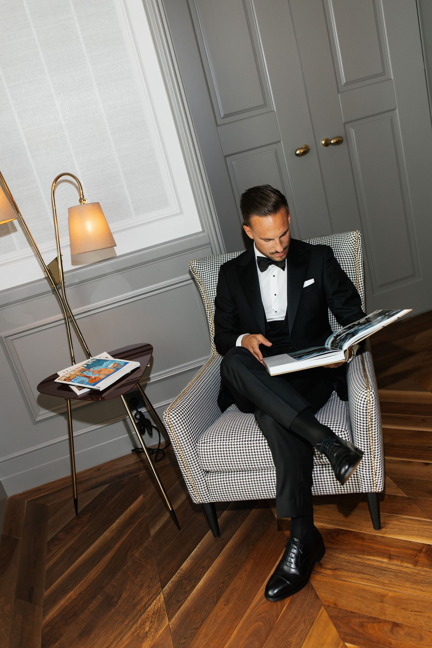 Dapper groom in black tuxedo reading a book in a sophisticated gray-paneled hotel suite.