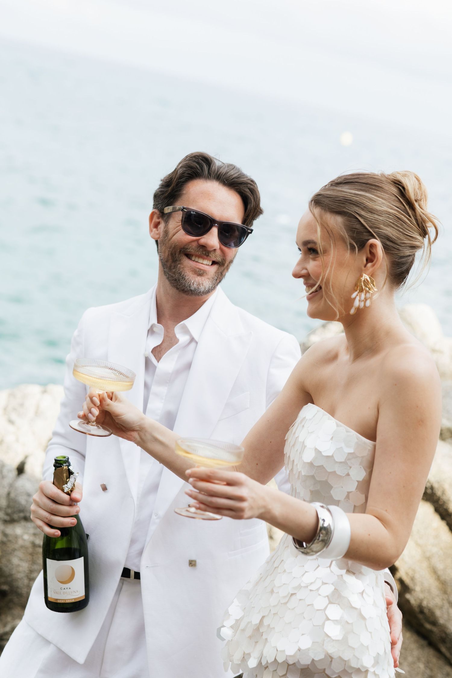 Elegant couple in white attire toasting with champagne glasses on rocky seaside with ocean backdrop.