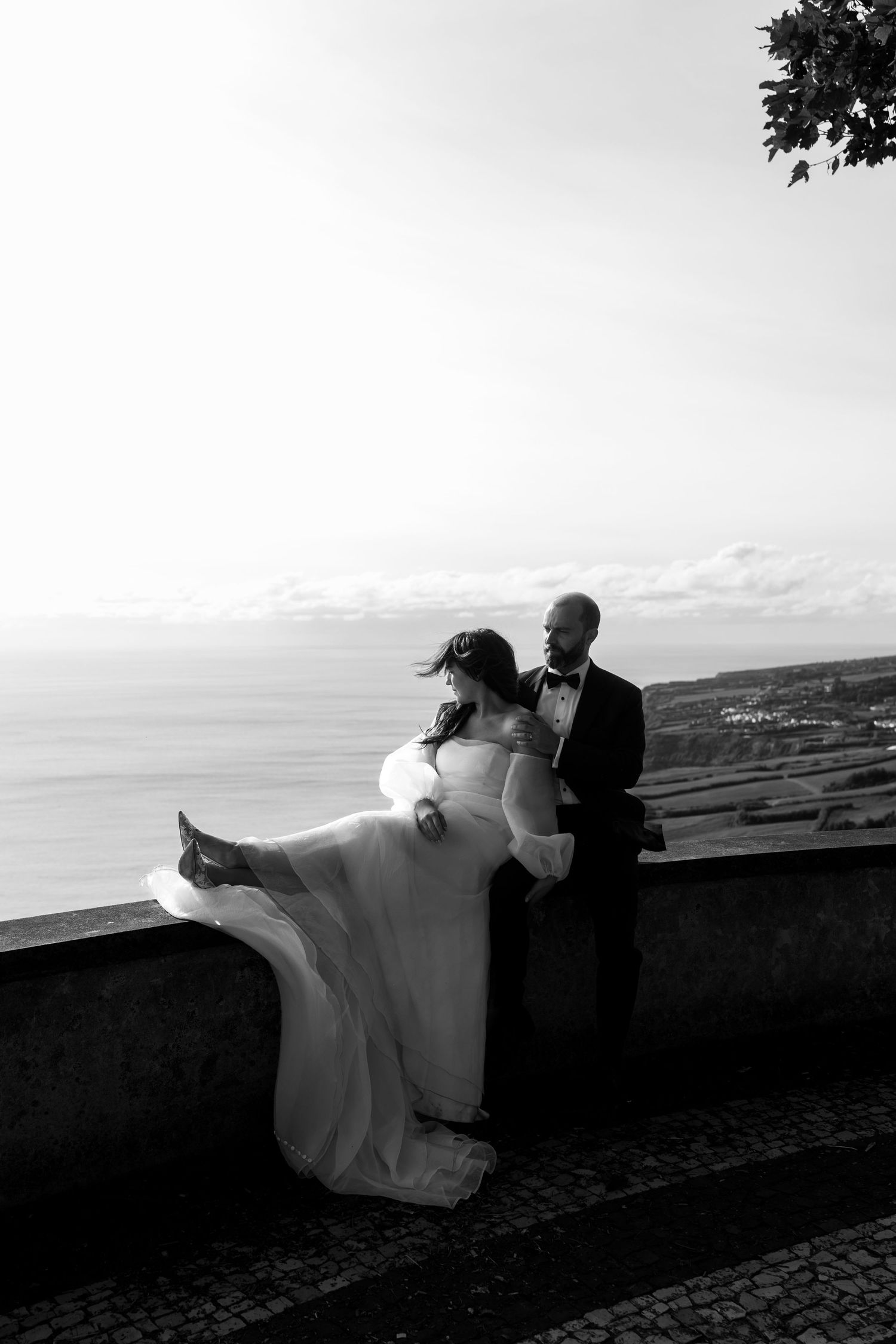 Black and white romantic wedding photo of bride and groom embracing by a stone wall overlooking the ocean.