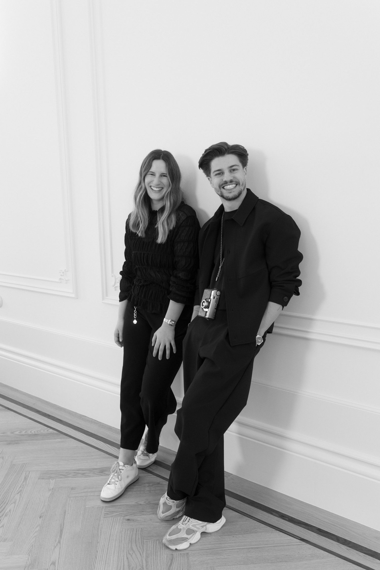 Black and white portrait of two smiling wedding photographers in all black outfits leaning against a wall.