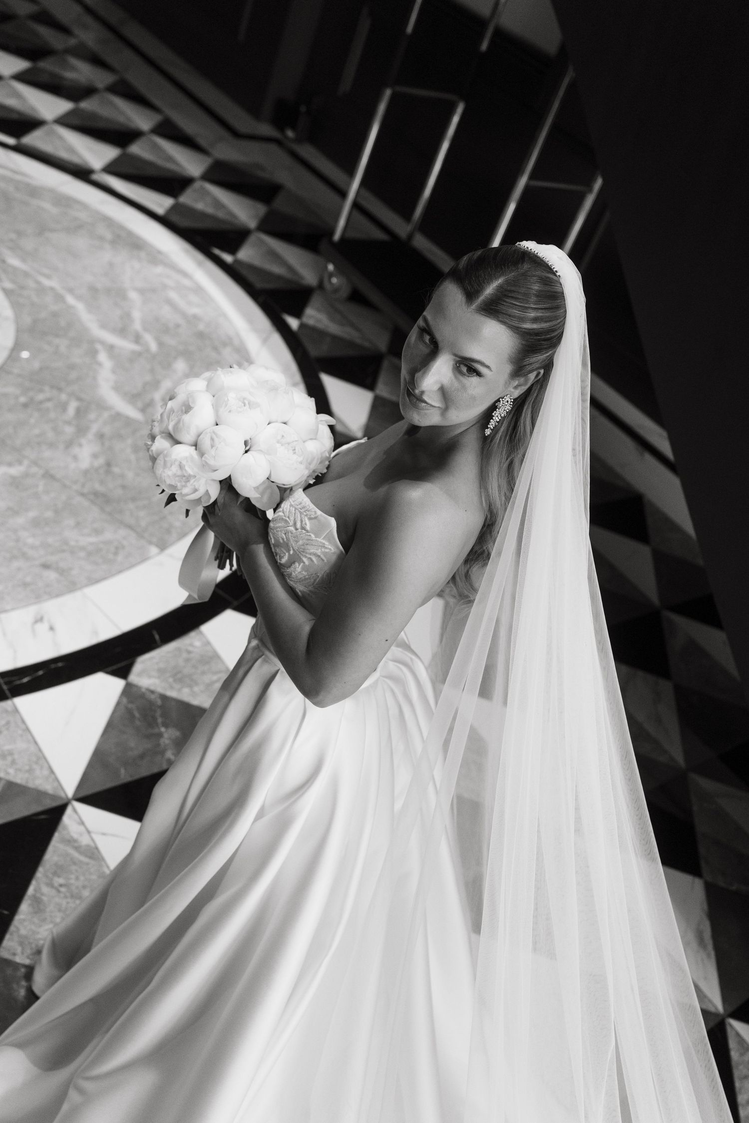 Elegant bride holding white peony bouquet in satin gown with long veil on grand staircase, black and white photo.