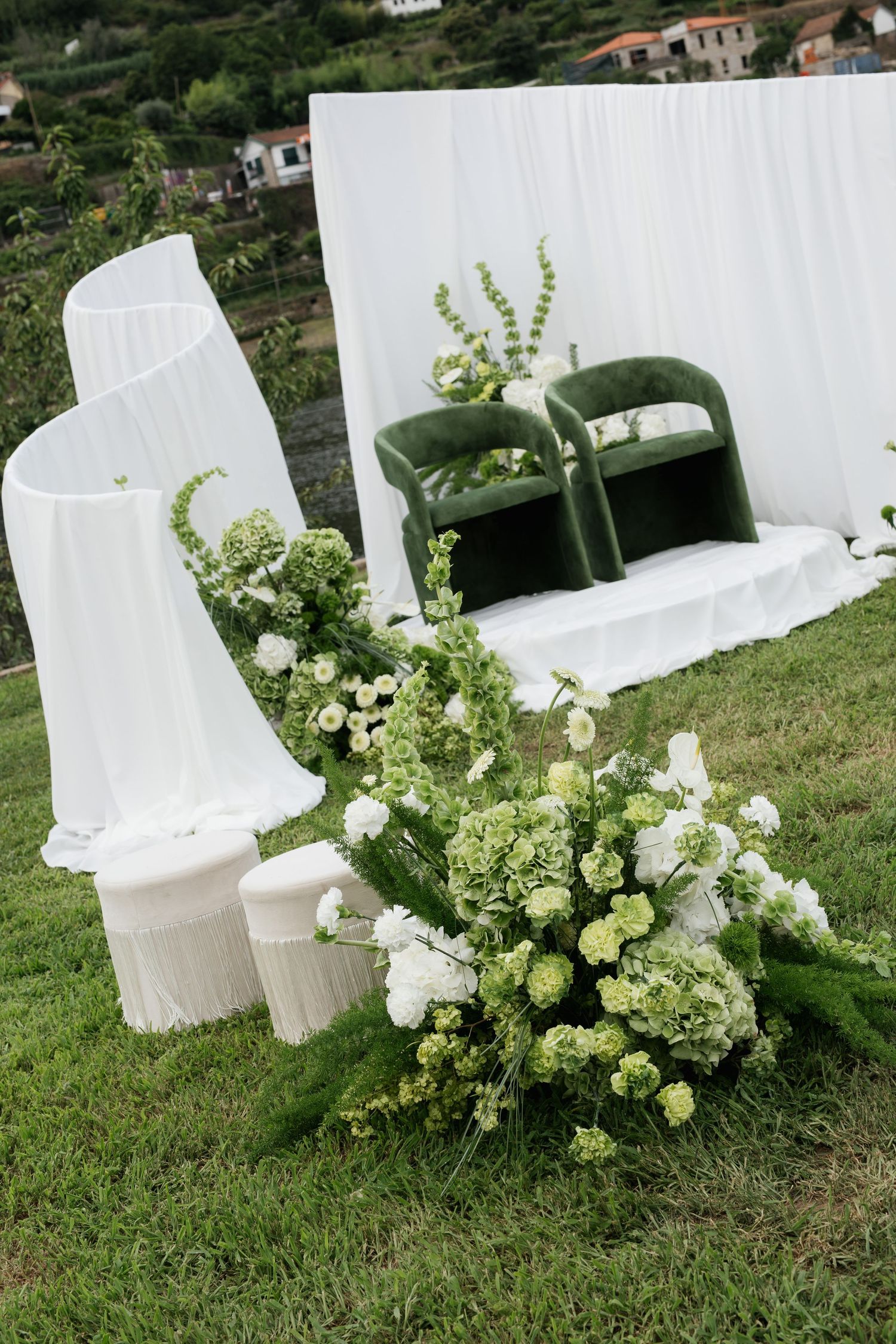 Elegant outdoor wedding ceremony setup with green velvet chairs, white draping, and lush green and white floral arrangements.