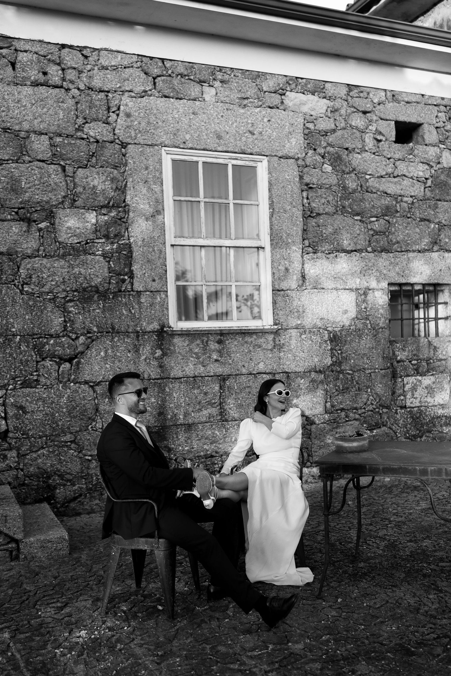 Black and white photo of bride and groom relaxing in chairs against a rustic stone wall.