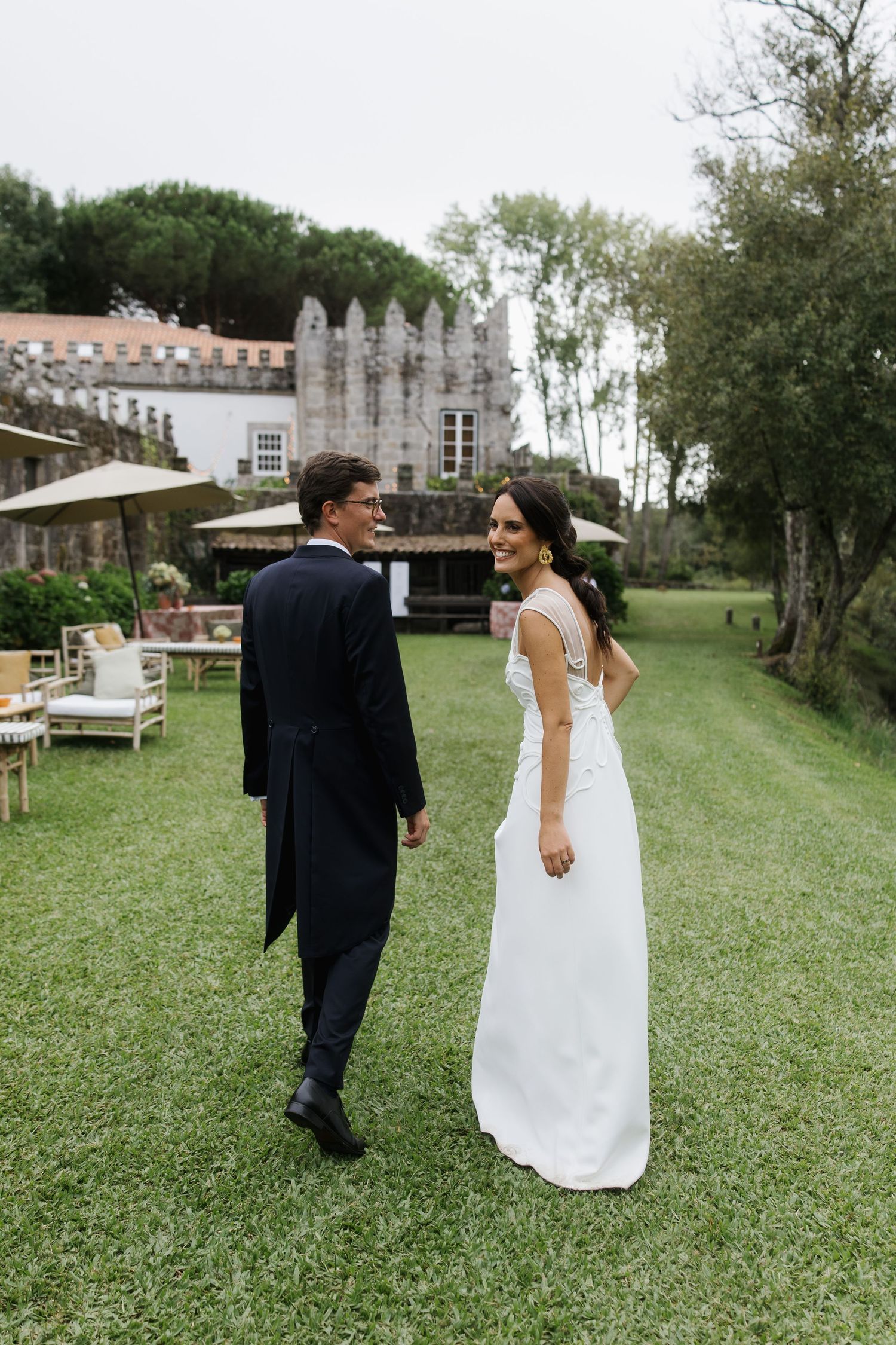 Bride in elegant white gown smiles at groom in dark suit while walking on green lawn near historic estate.