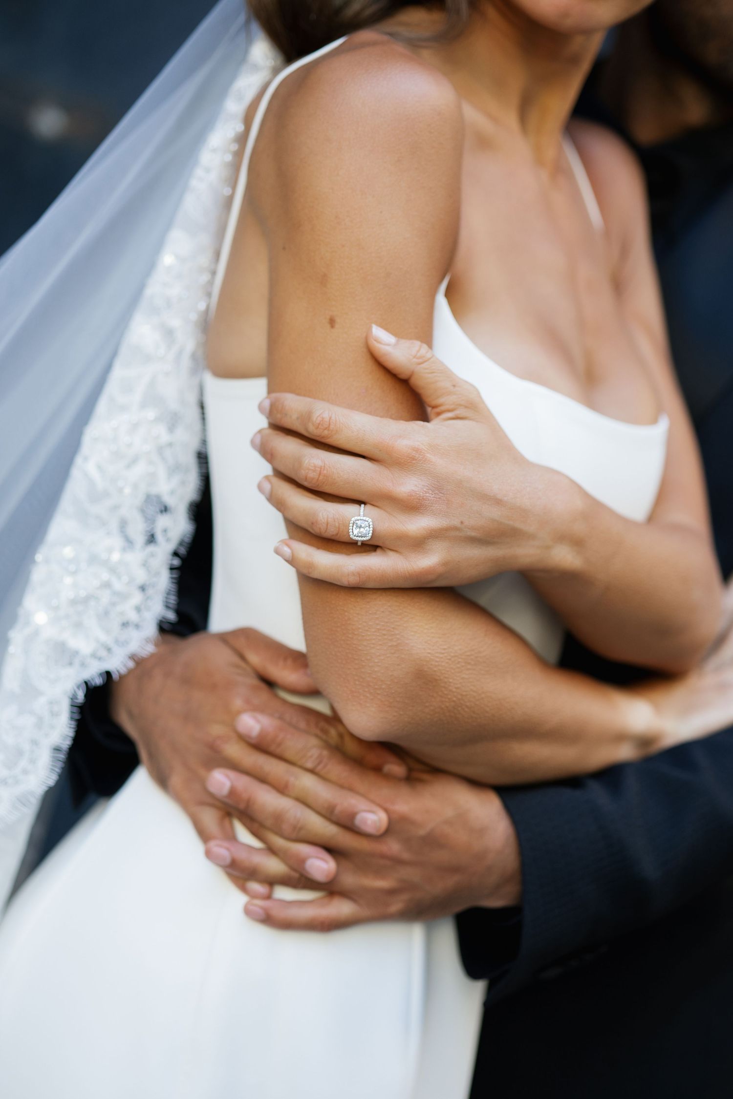 Close-up of bride wearing elegant diamond engagement ring while groom embraces her in wedding attire.
