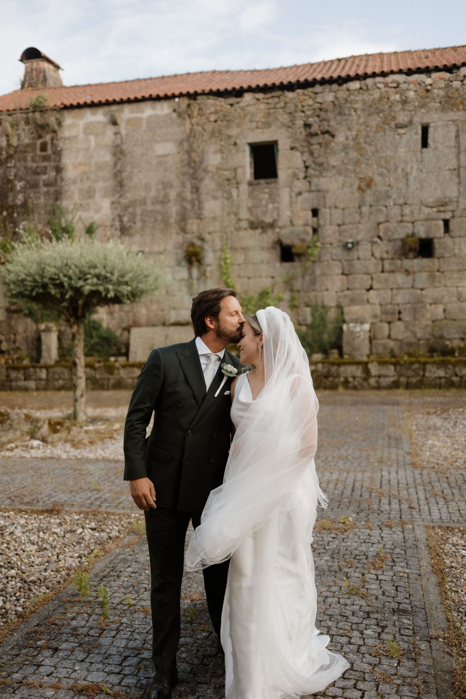 Bride and groom sharing a tender kiss in a cobblestone courtyard beside a historic stone building.