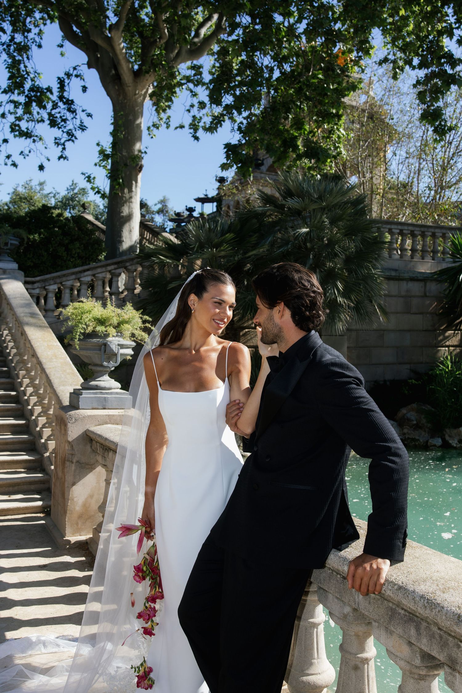 Bride and groom share a romantic moment on stone bridge steps surrounded by lush gardens and turquoise water.