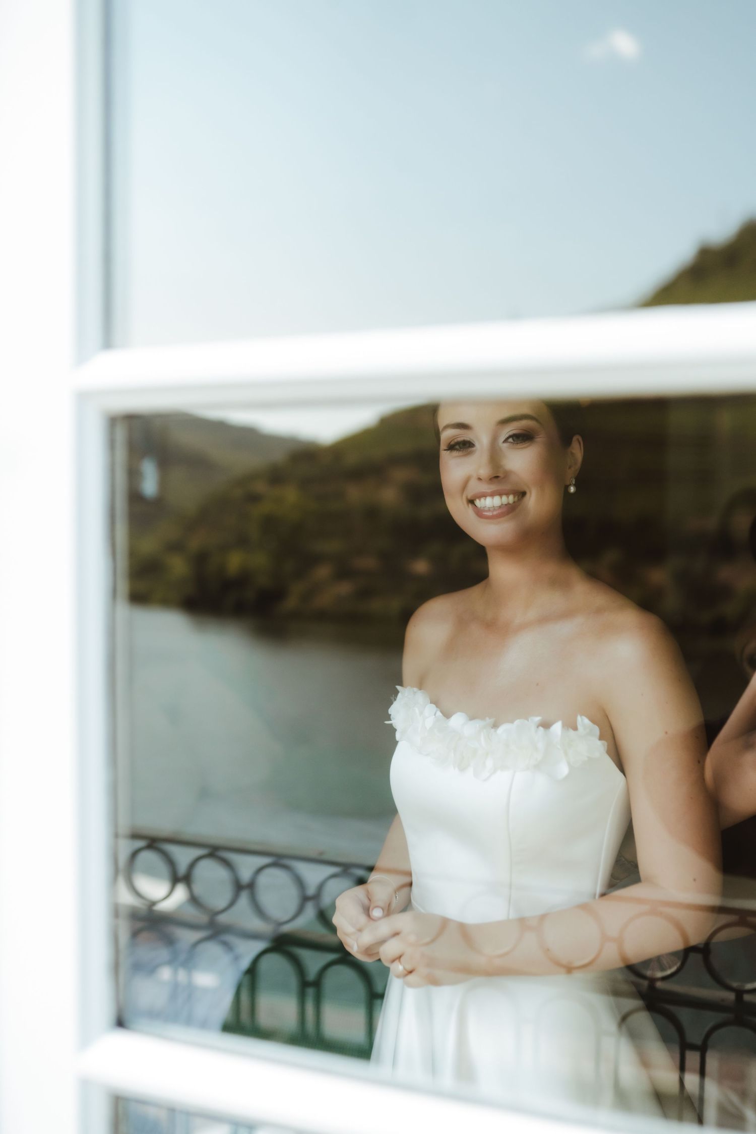 Smiling bride in strapless white gown poses by a window with scenic mountain and river views behind her.