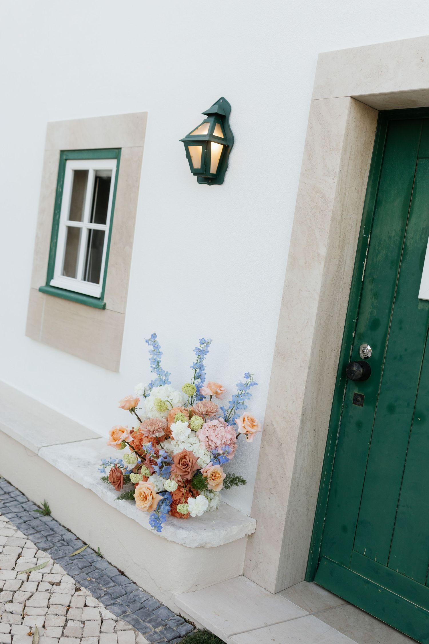 Colorful floral arrangement with peach roses and blue delphiniums displayed beside green door of white venue.