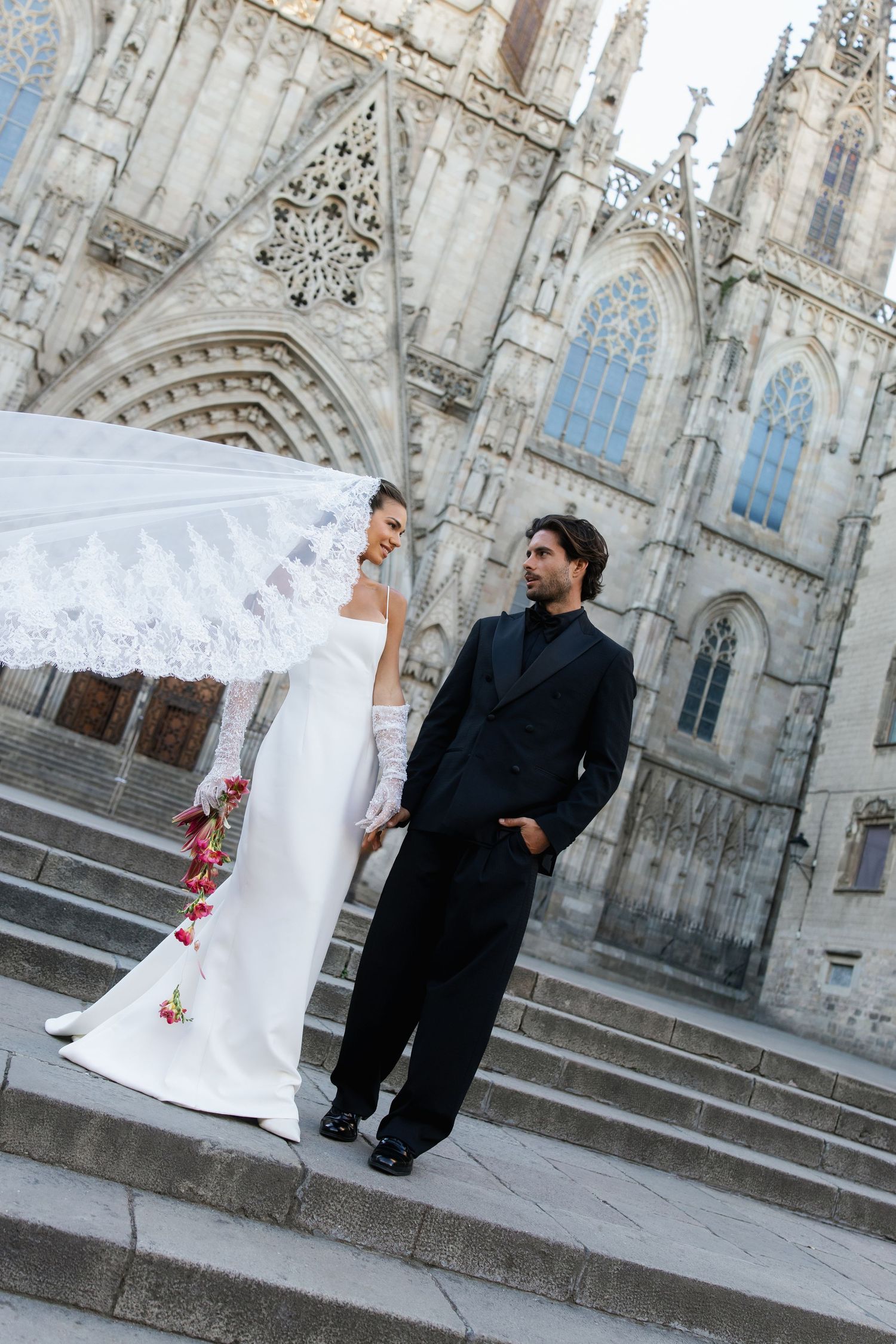 Bride in white gown with long lace veil and groom in black suit posing on cathedral steps in Barcelona.