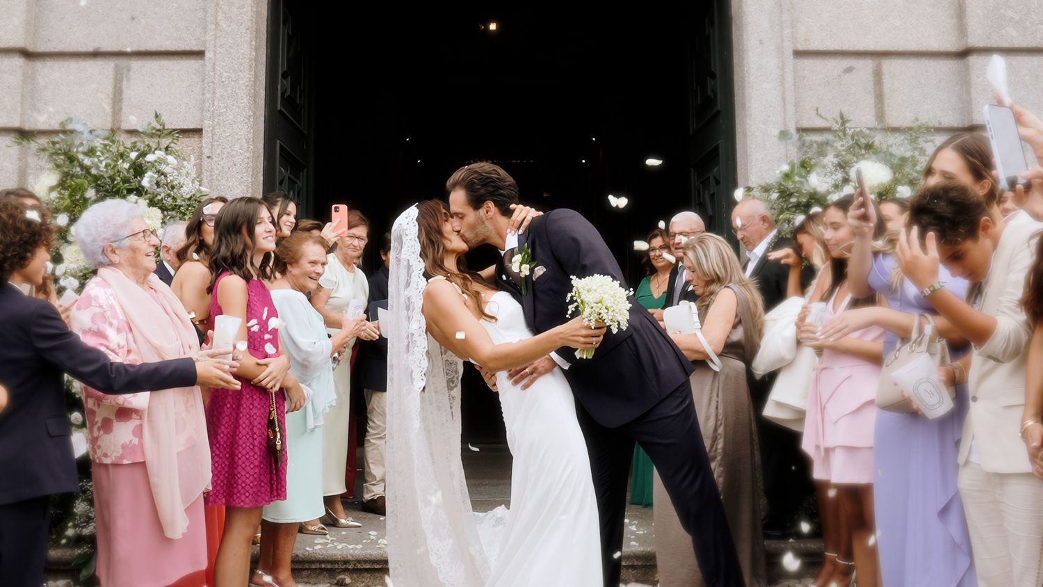 Newlyweds share a kiss outside a church entrance as guests celebrate with confetti and applause.