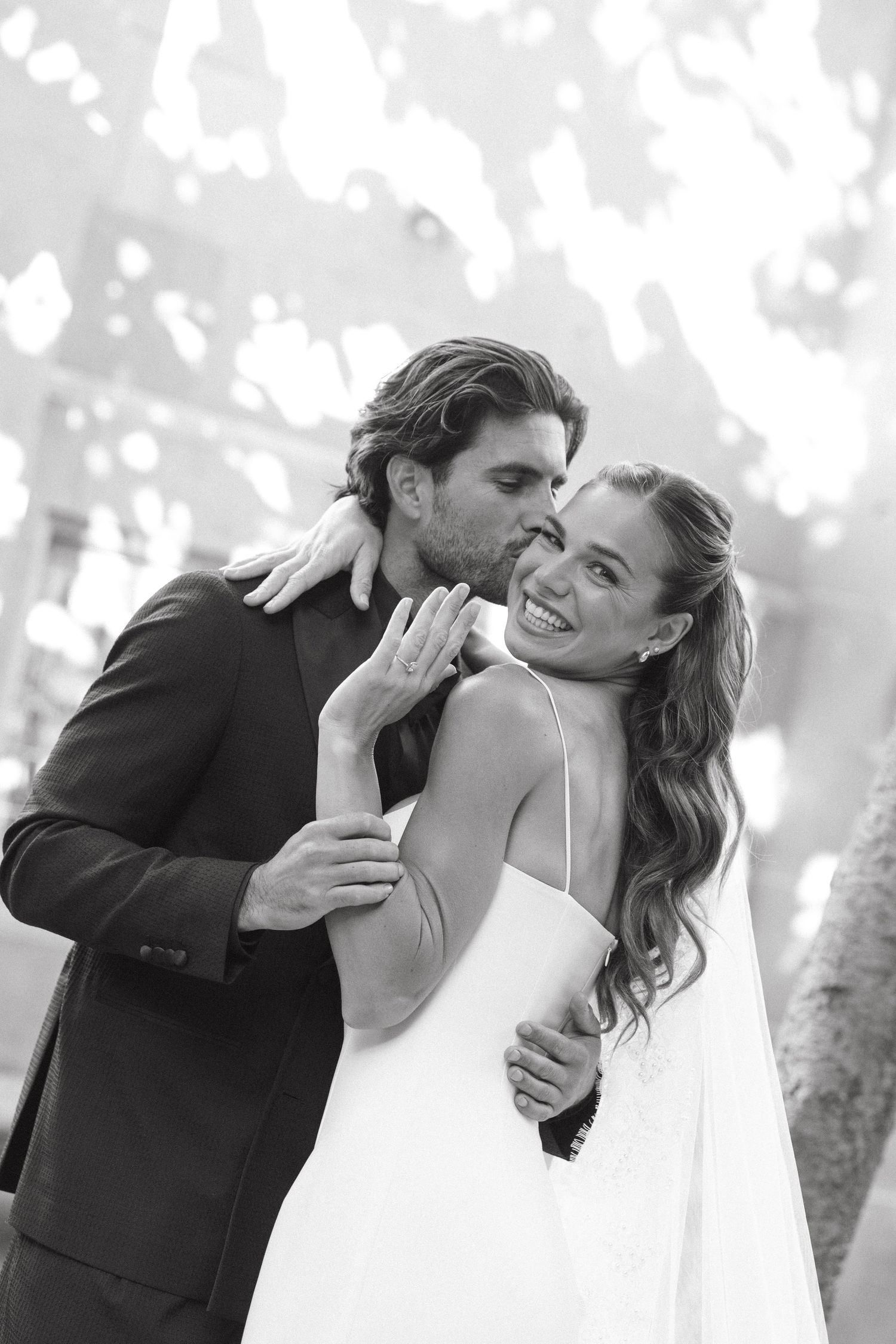 Black and white photo of groom kissing smiling bride on cheek during romantic wedding celebration.