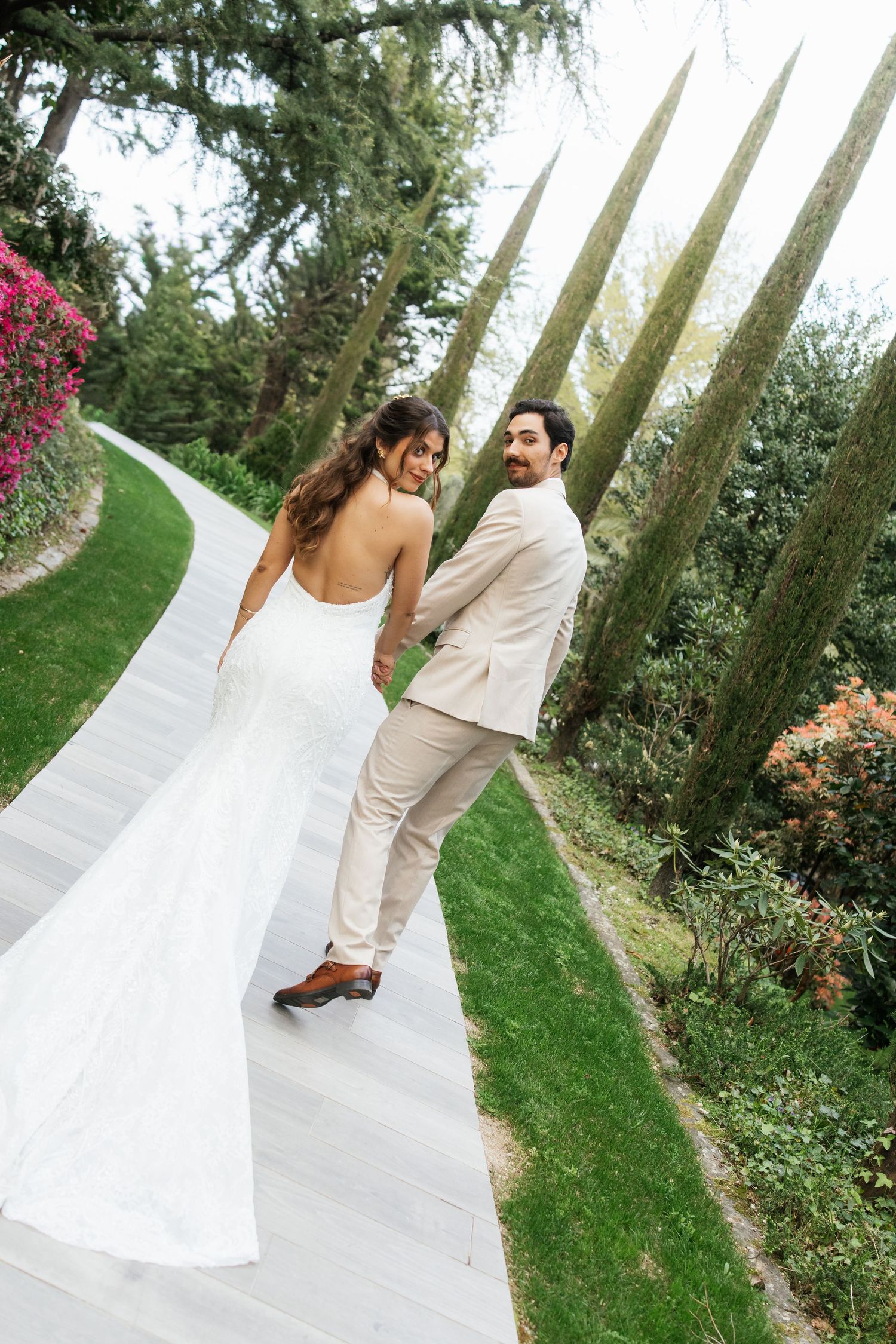 Bride in backless white gown and groom in beige suit walk hand-in-hand through a lush garden path.
