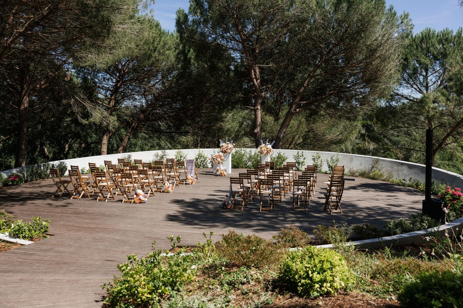 Outdoor wedding ceremony setup with wooden chairs arranged in semicircle on deck surrounded by pine trees.