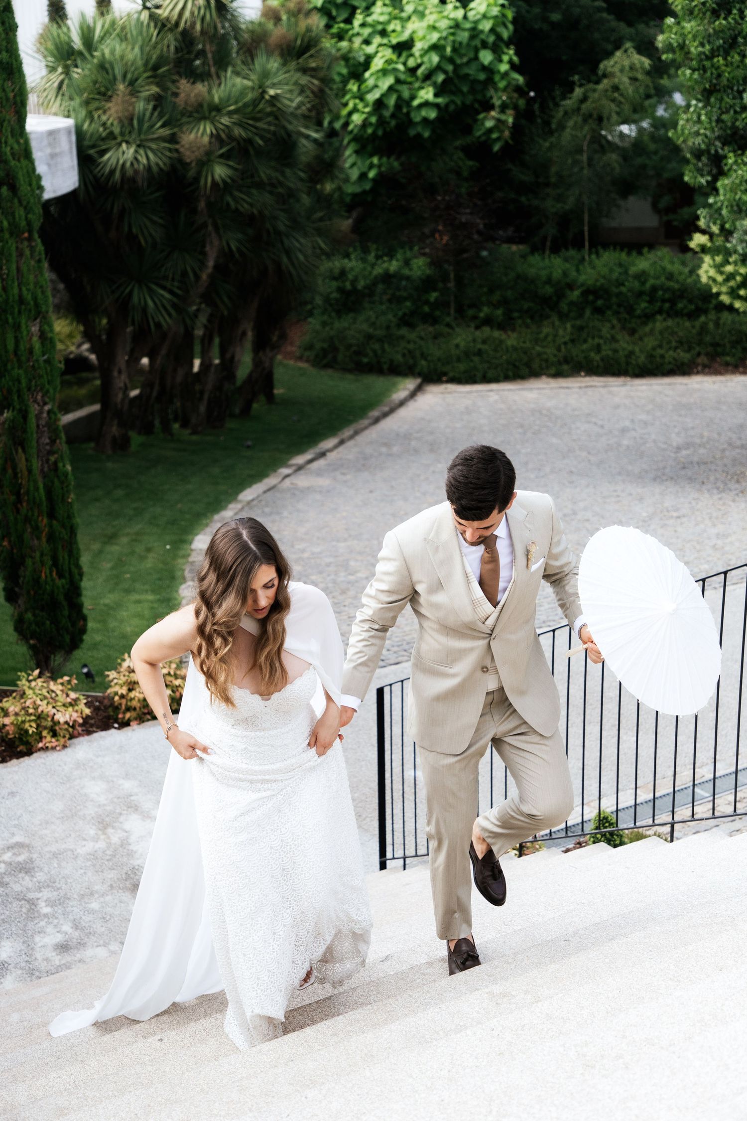 Bride in flowing white gown and groom in beige suit ascending outdoor stone steps in a lush garden.