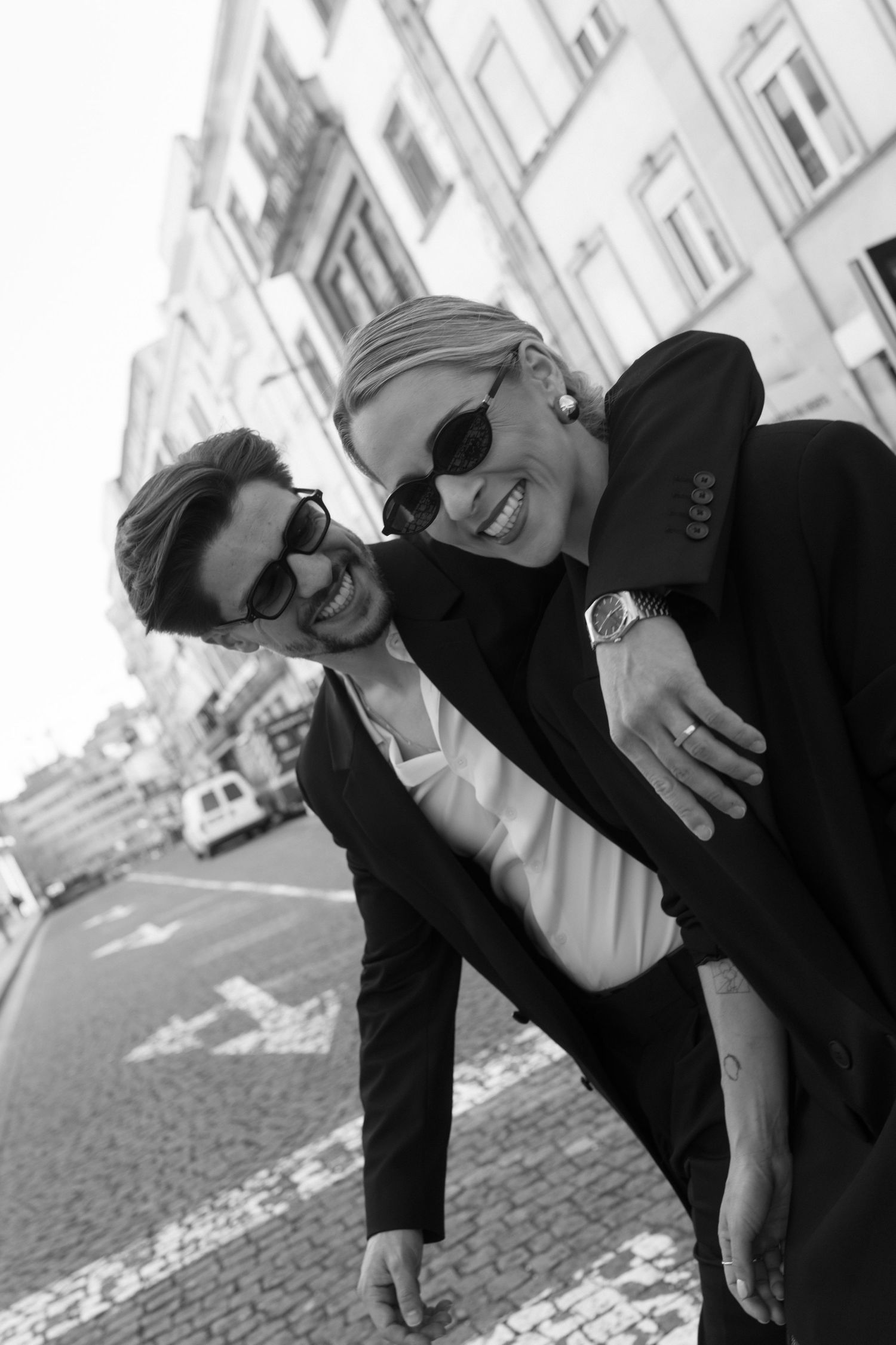 Black and white photo of laughing couple in black suits embracing on a cobblestone city street.