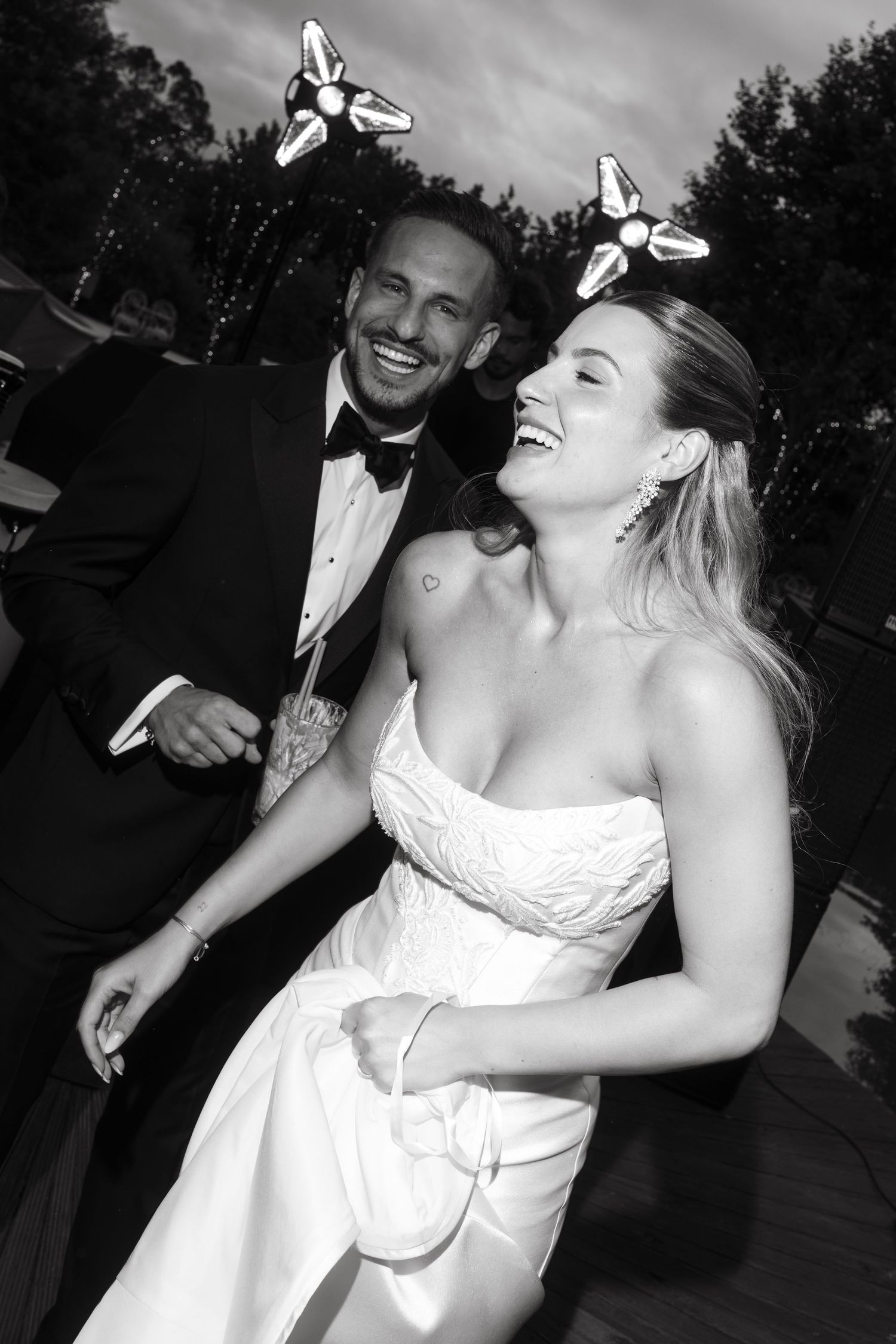 Black and white photo of a laughing bride in strapless gown and groom in tuxedo wearing novelty headbands at wedding reception.