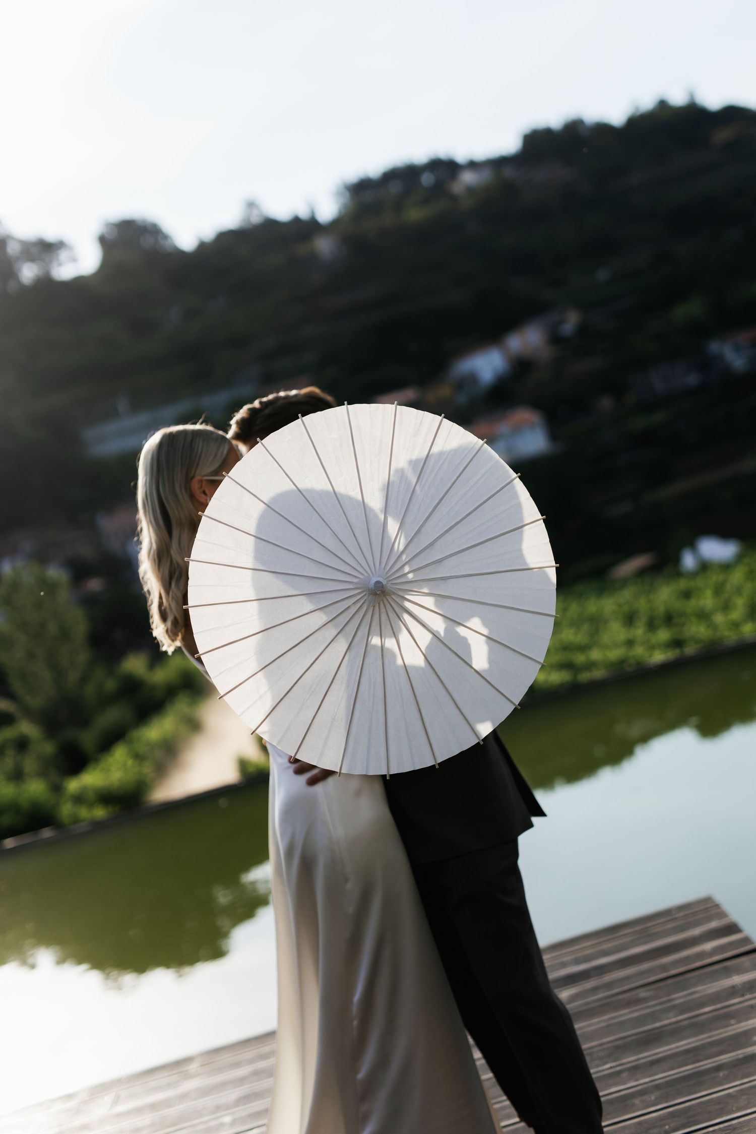 Bride and groom share romantic moment hidden behind white parasol on wooden dock with scenic lake backdrop.