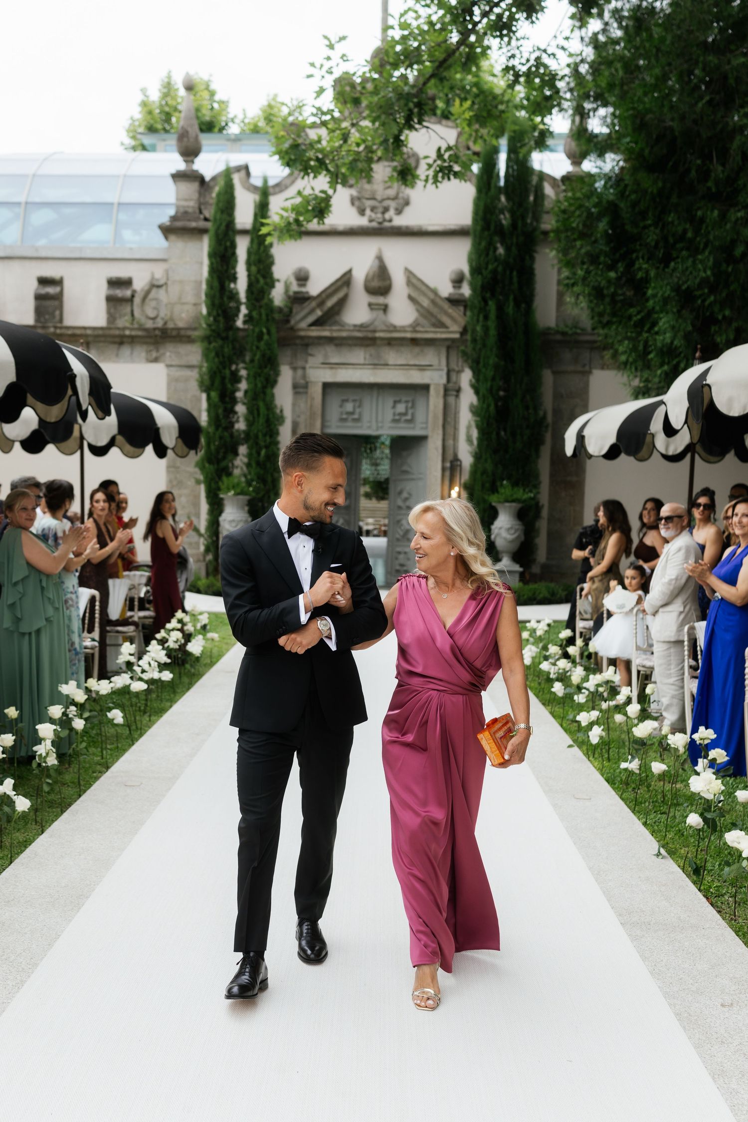 Groom in tuxedo walks down white aisle with woman in magenta jumpsuit at elegant outdoor wedding ceremony.