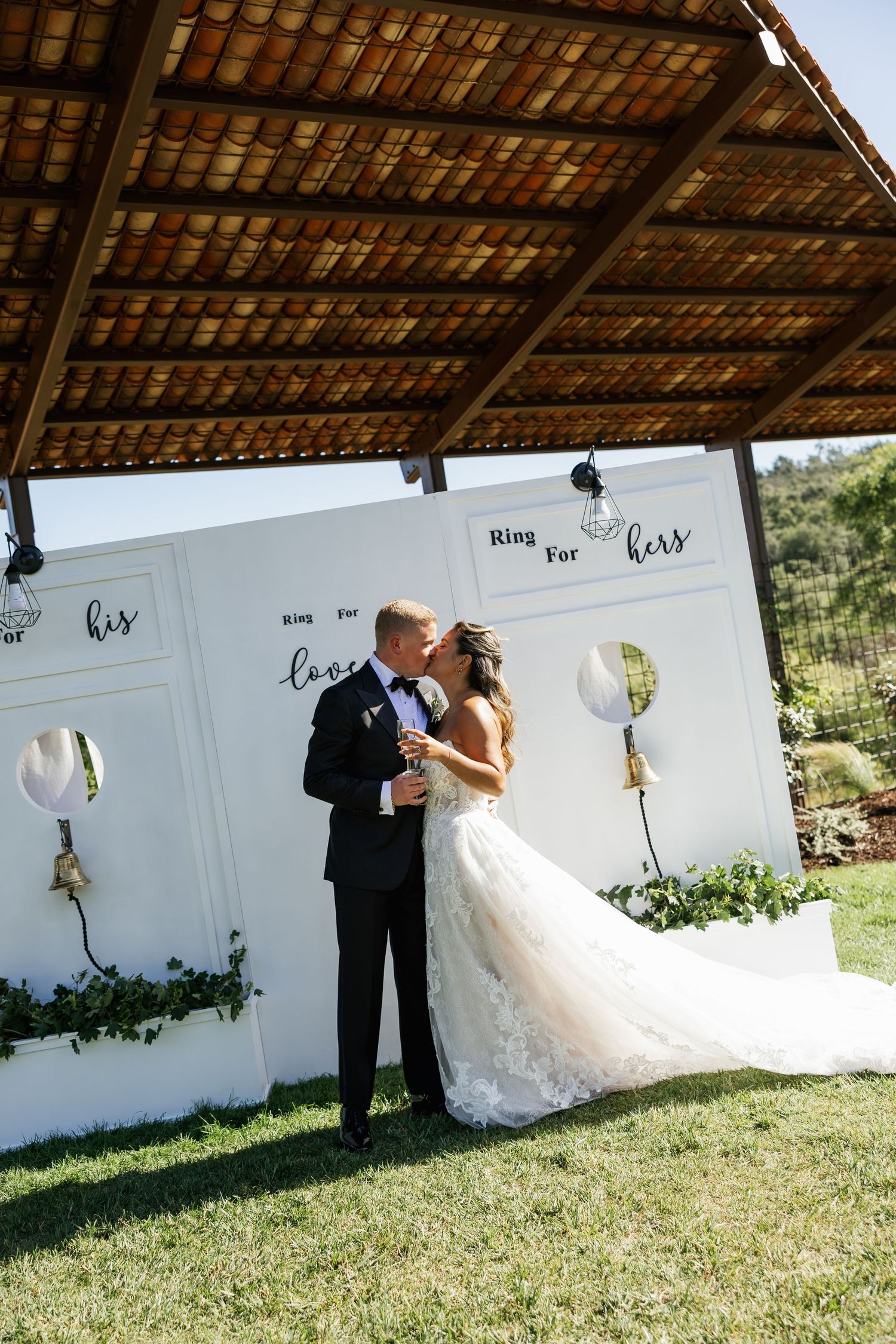 Bride and groom share a kiss at outdoor wedding ceremony in front of white panel backdrop with his and hers signage.