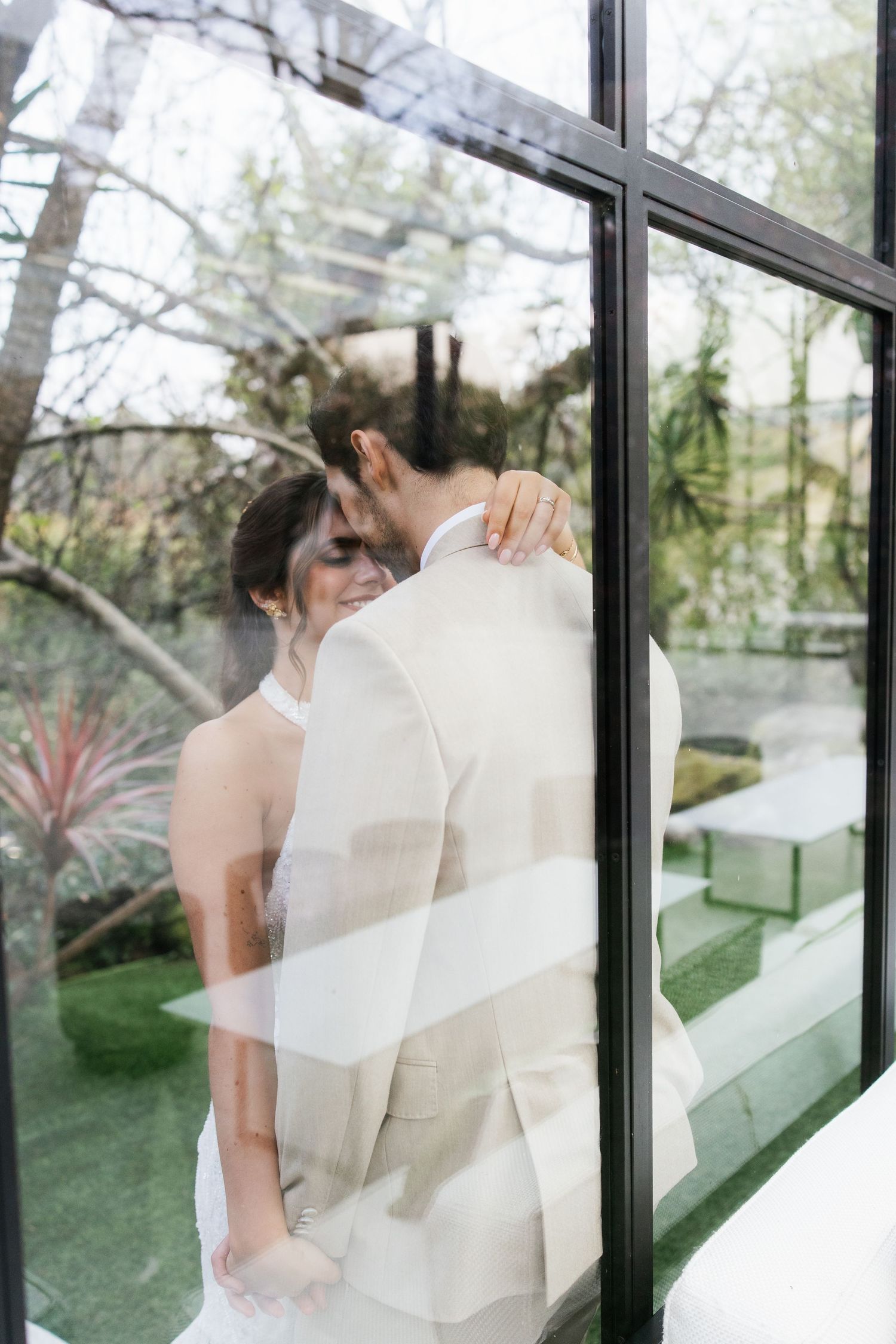 Romantic wedding portrait of bride and groom embracing, photographed through a glass window reflection.