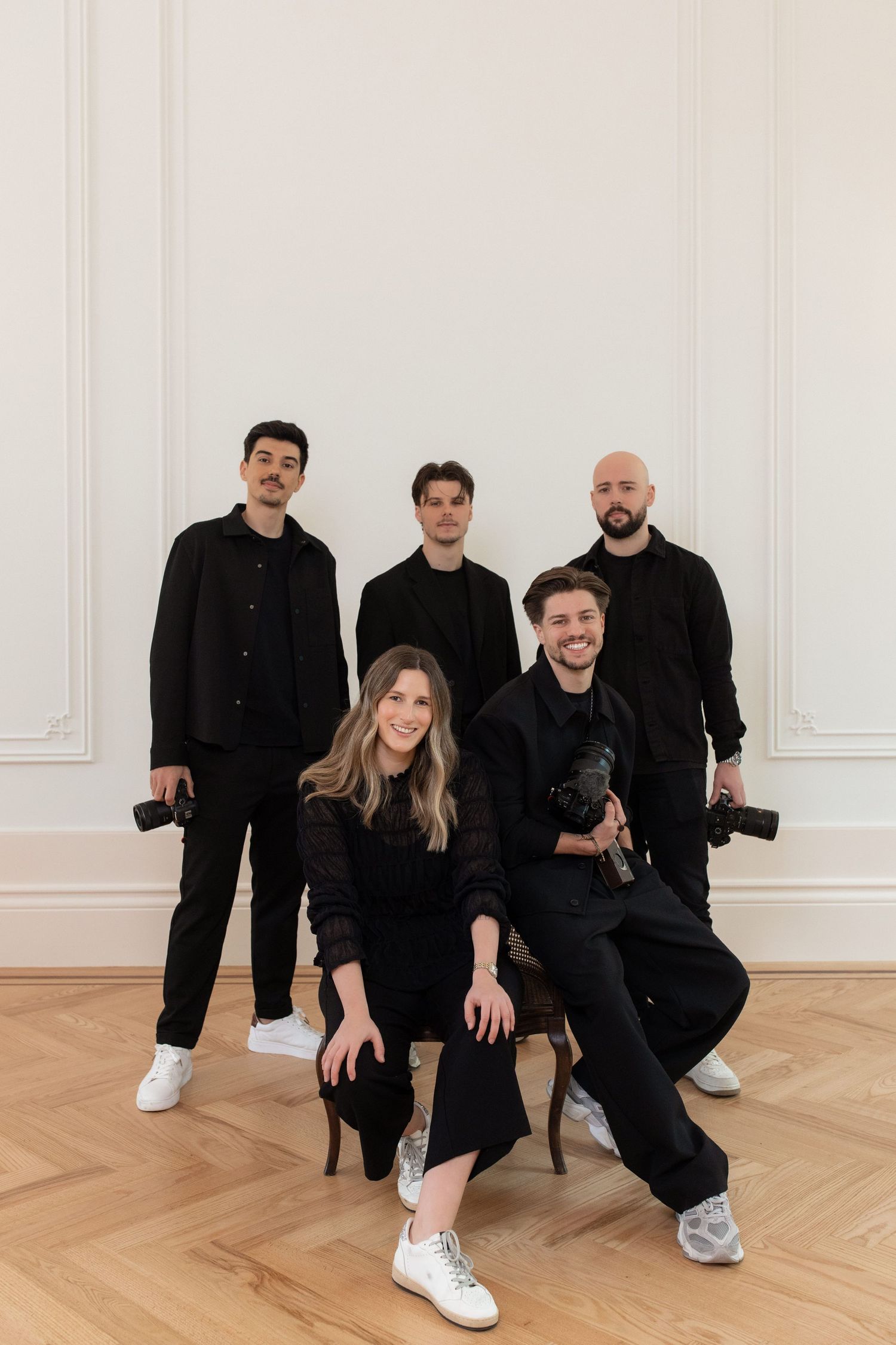 Professional wedding photography team of five dressed in black posing with cameras in an elegant white room.