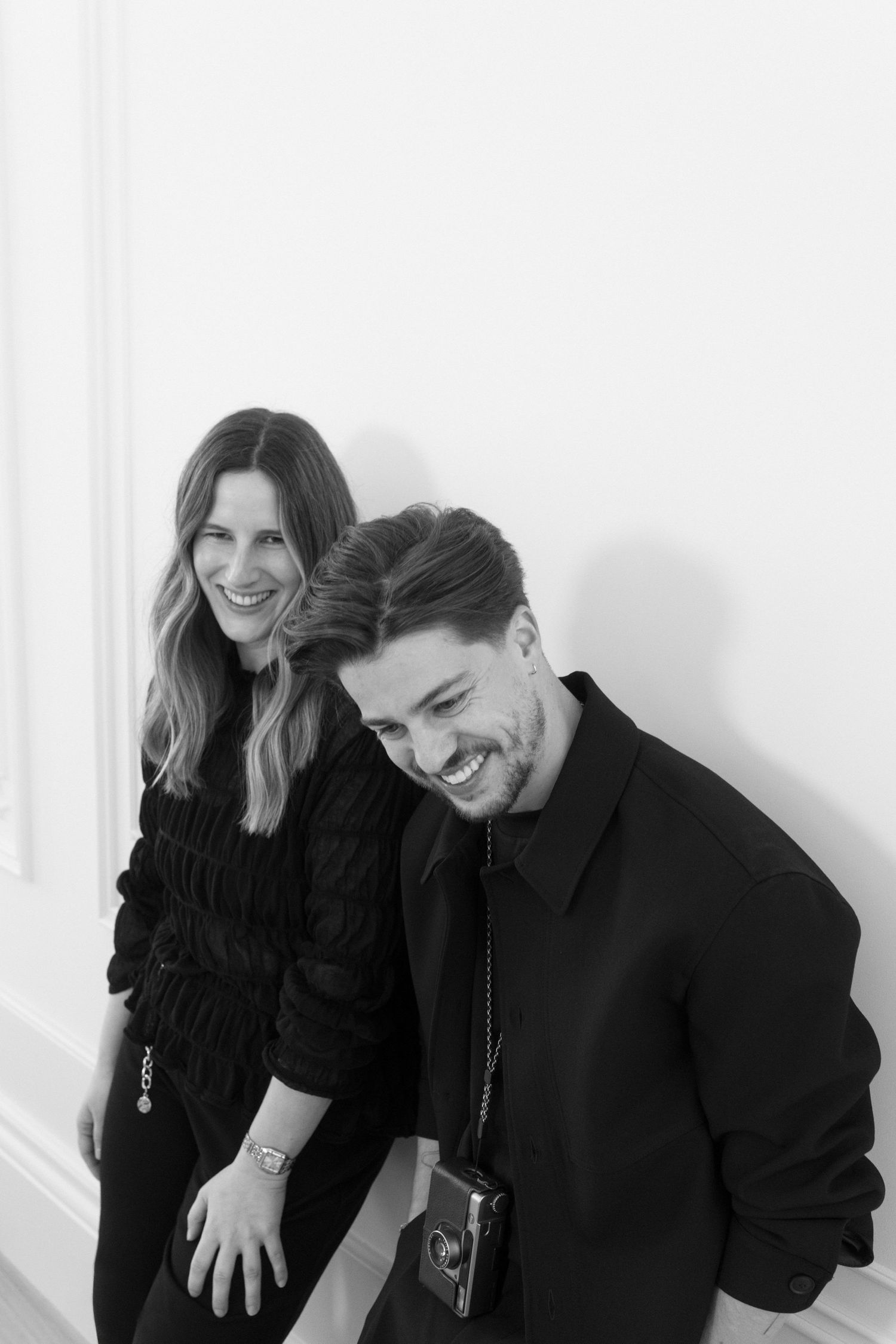 Black and white portrait of a smiling couple in black outfits leaning against a white wall.