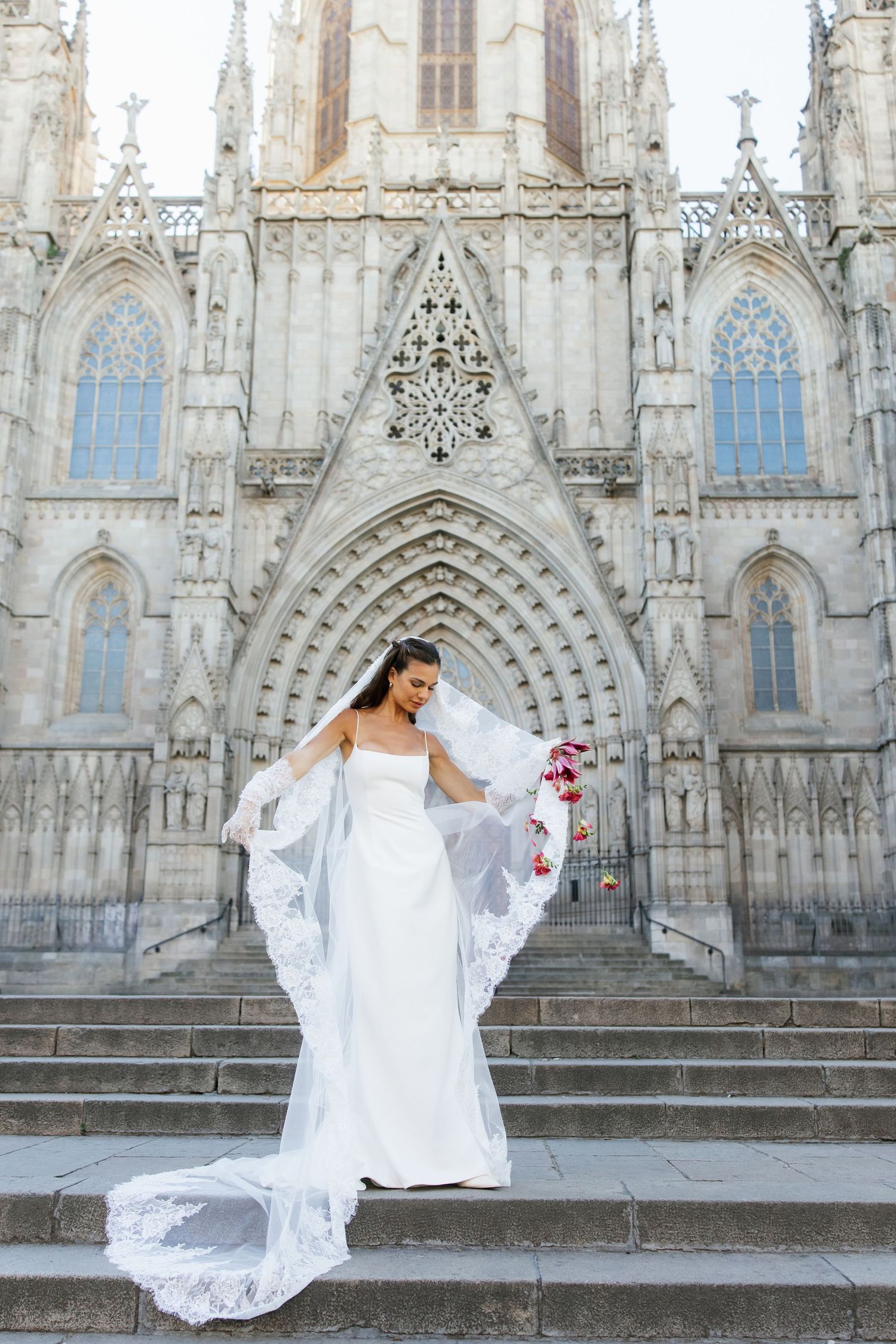 Bride in strapless white gown with lace veil posing on cathedral steps holding a bouquet of red flowers.