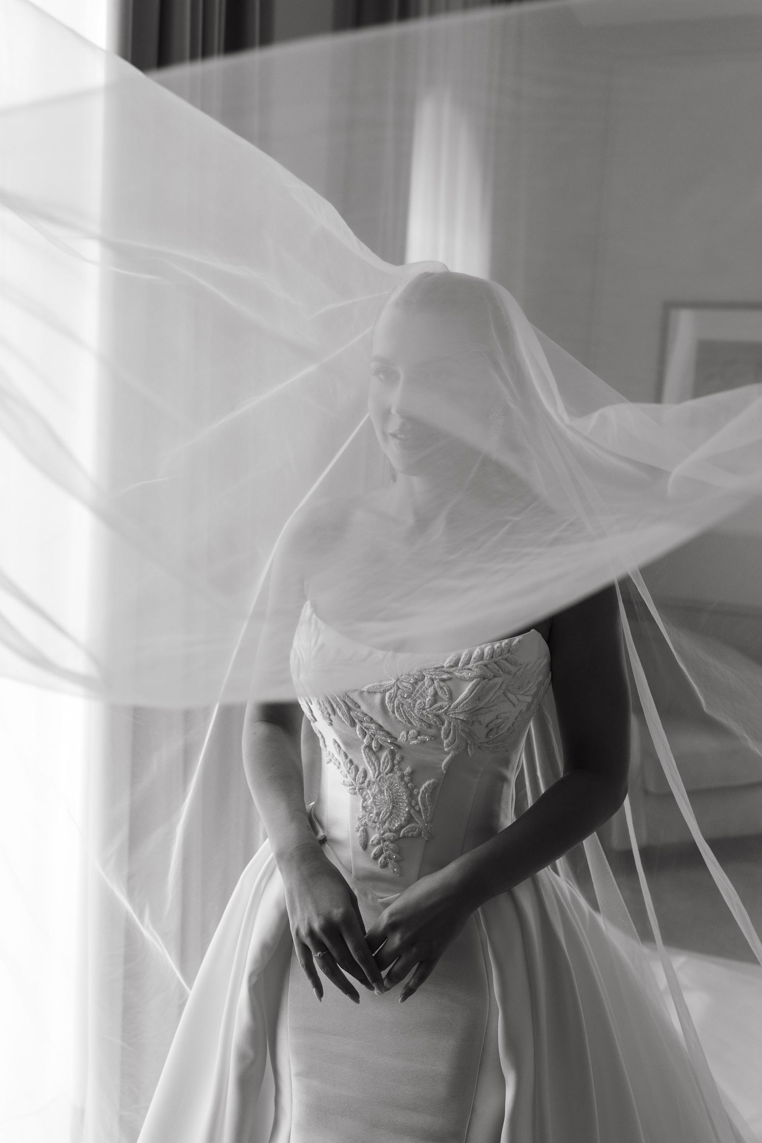 Bride in strapless lace wedding gown with flowing veil billowing dramatically in black and white portrait.