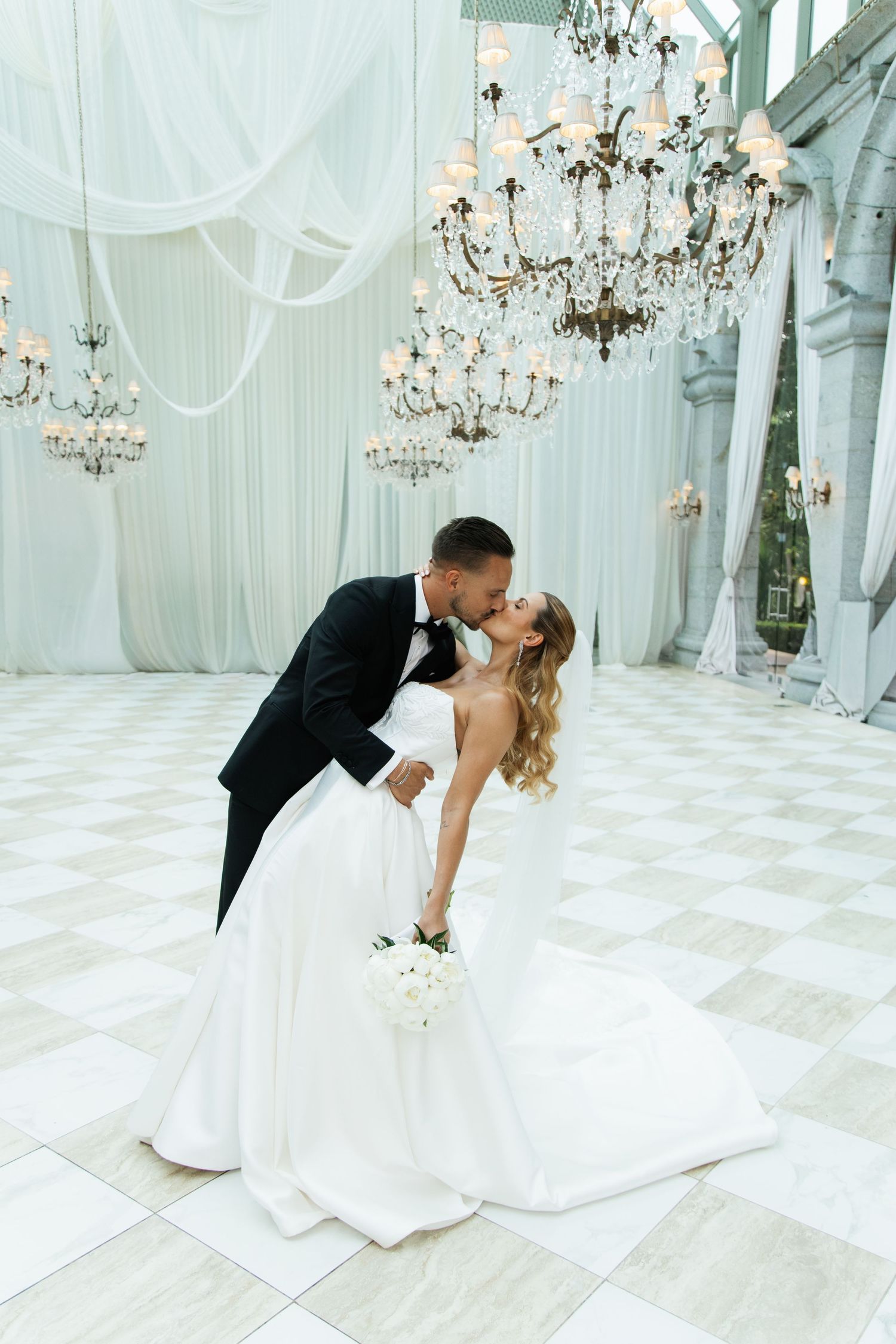 A bride and groom share a romantic dip kiss on a white marble dance floor beneath elegant crystal chandeliers.