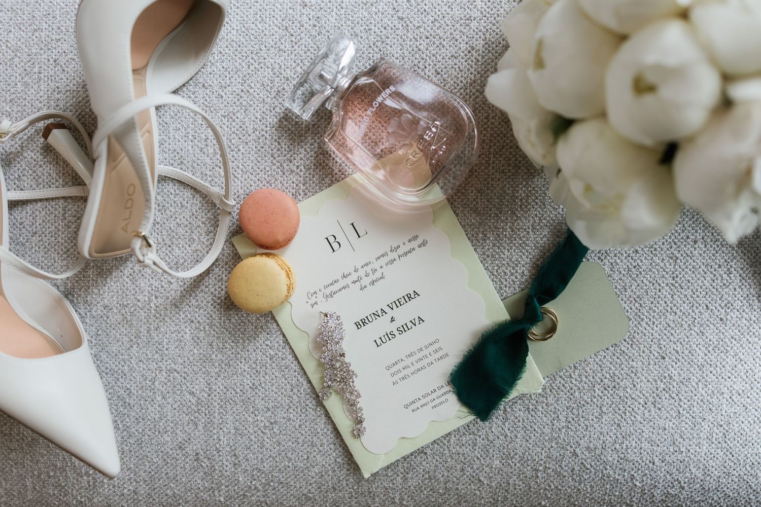 Elegant wedding flat lay featuring invitation, heels, macarons, perfume, and floral bouquet on gray fabric.