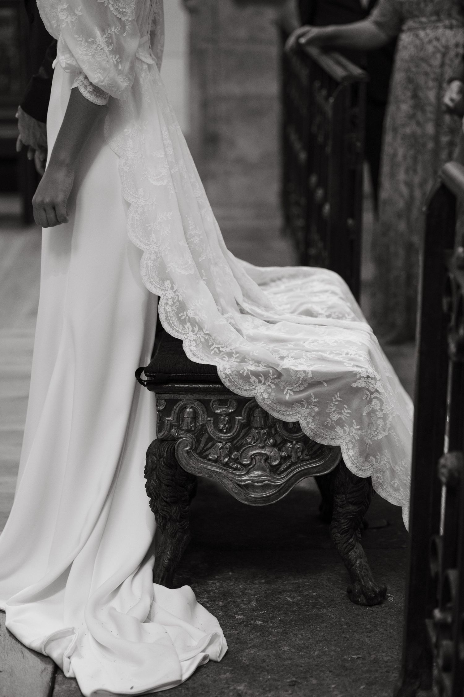 Black and white close-up of bride in lace wedding gown with elegant train draped over ornate wooden bench.