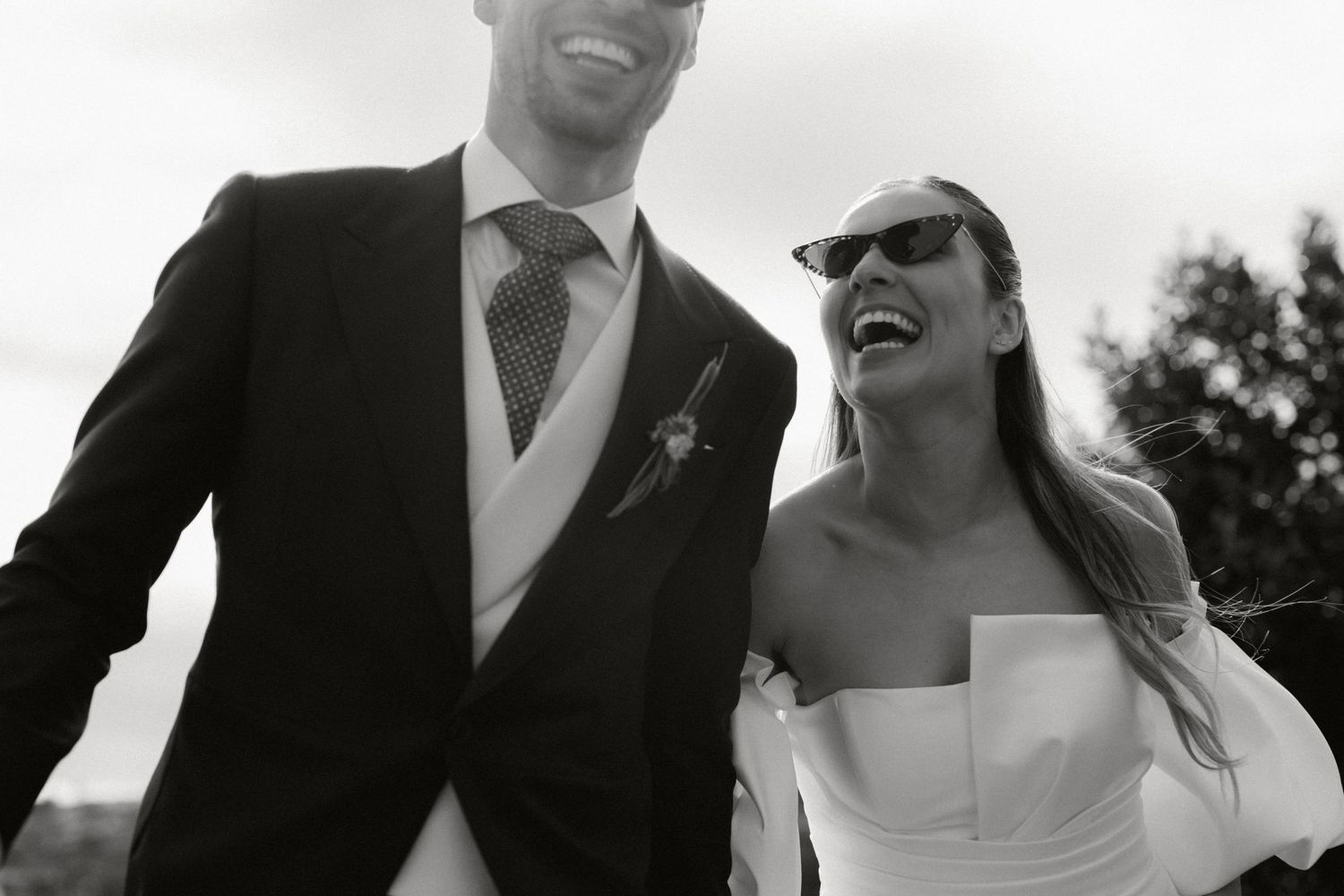 Black and white photo of laughing bride in strapless gown and groom in dark suit walking together outdoors.
