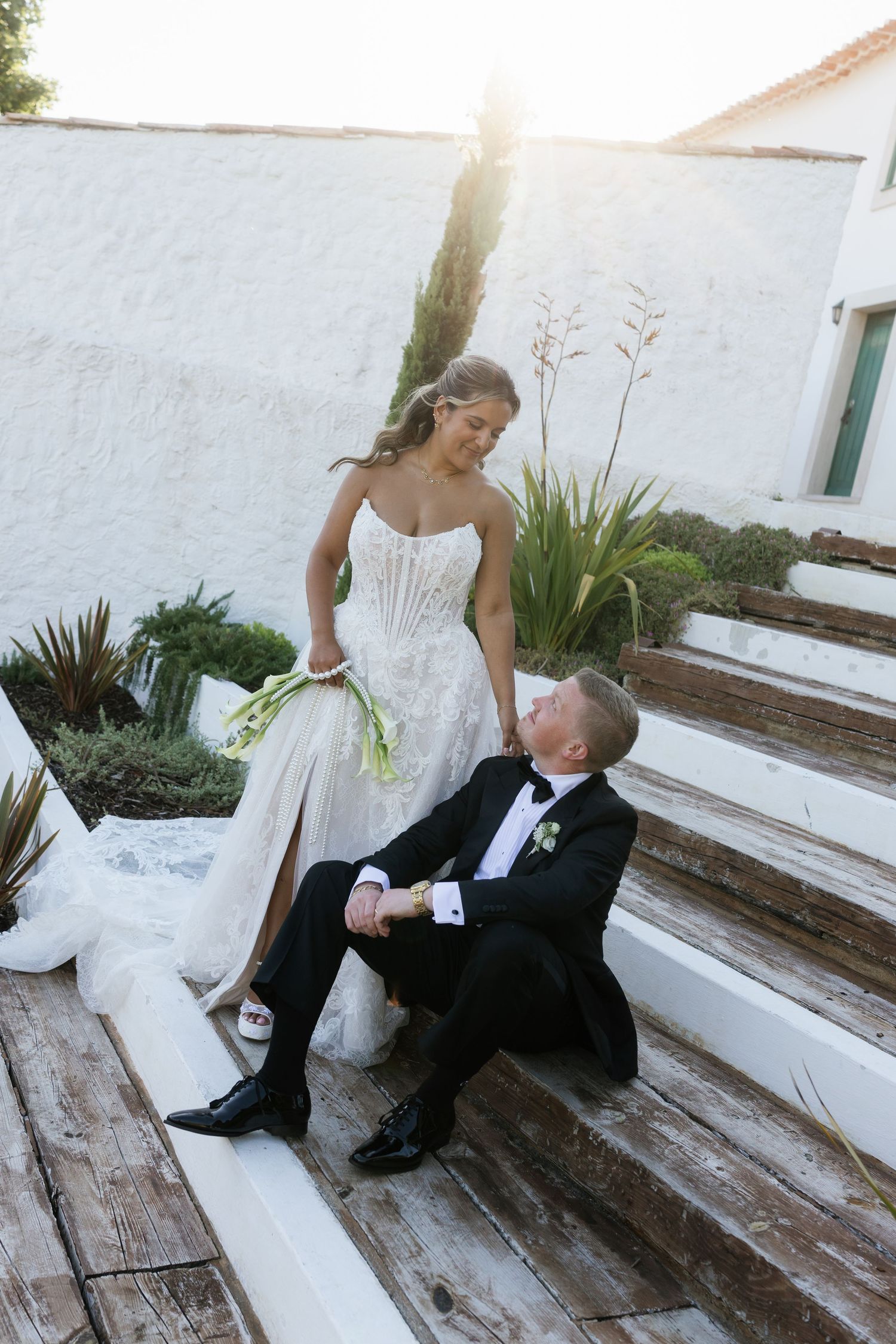 Groom gazes up adoringly at bride in corset lace gown on sunlit wooden steps at outdoor wedding venue.