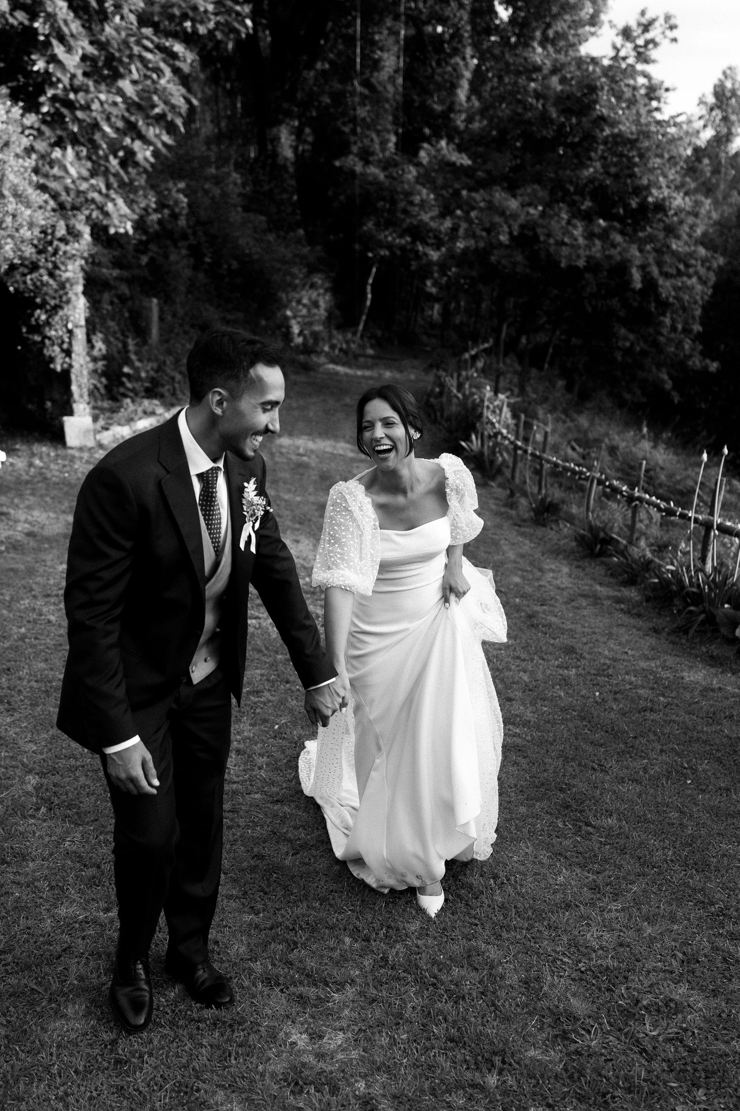 Black and white photo of a laughing bride and groom walking hand in hand through a lush garden path.