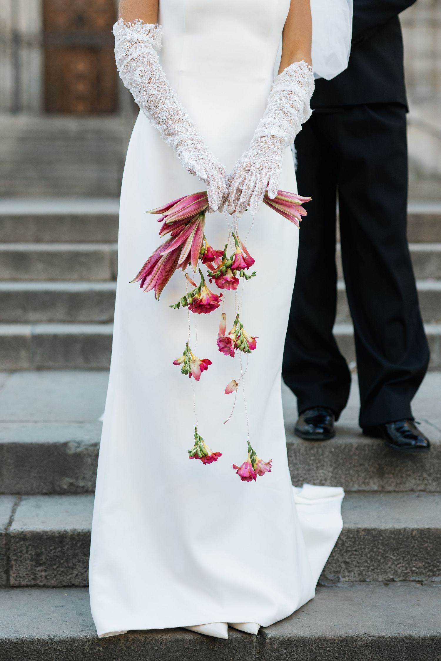Bride in white gown and lace gloves holds cascading pink tulip and rose bouquet on stone steps.