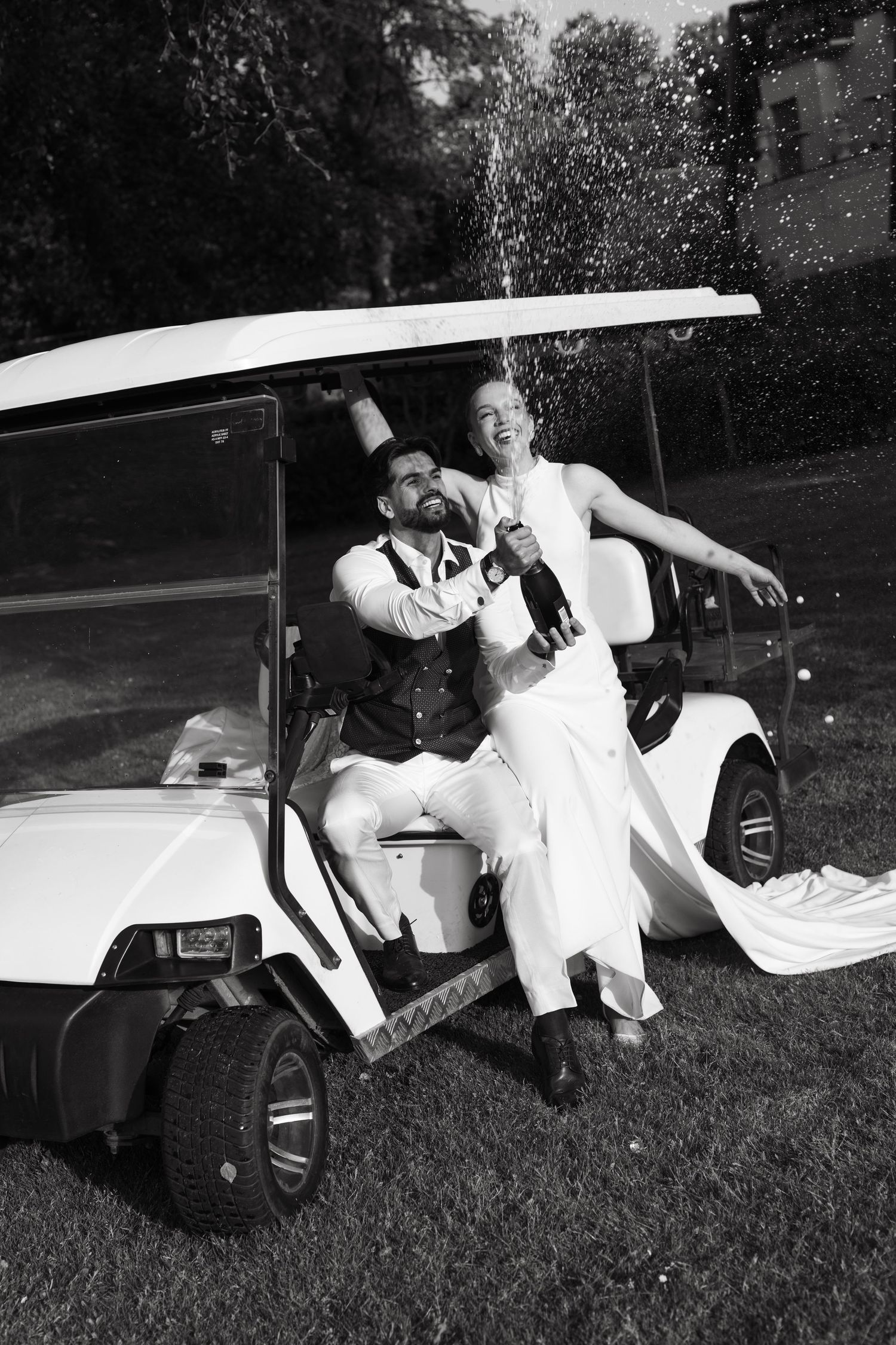 Black and white photo of joyful newlyweds popping champagne bottle while sitting on white golf cart.